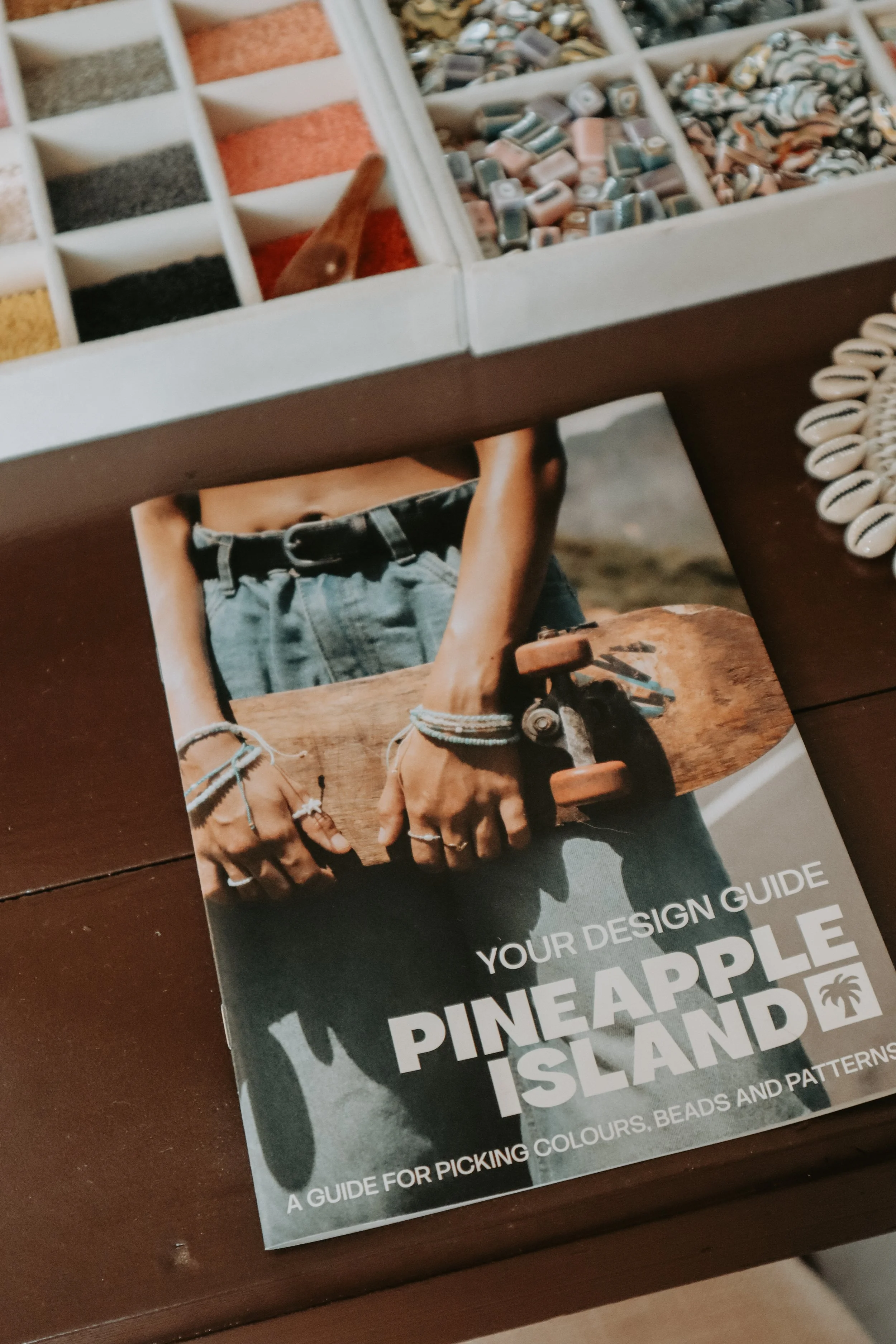 A guidebook titled 'Your Design Guide for Picking Colours, Beads and Patterns' features a person holding a wooden object, wearing multiple bracelets, and dressed in denim shorts.