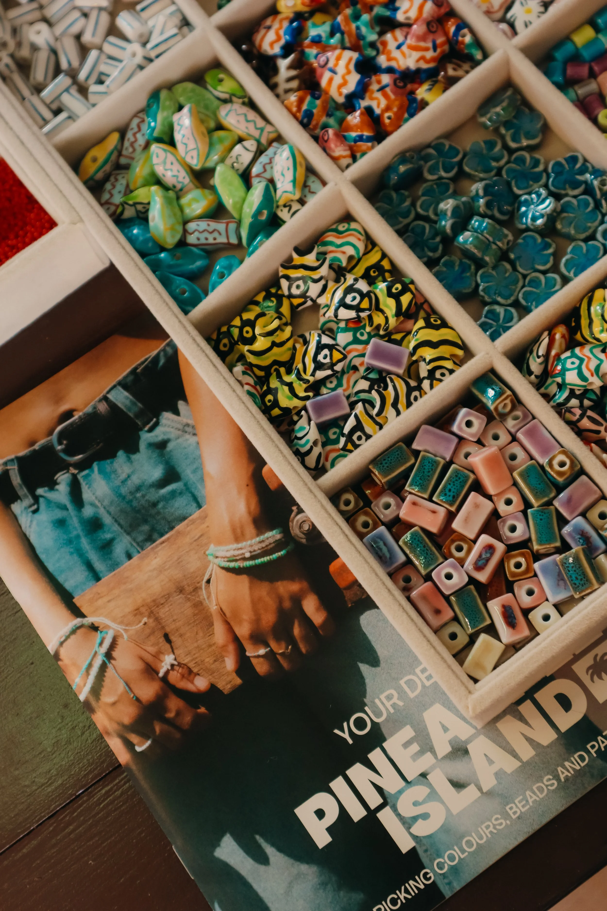Close-up of a compartmentalized box containing colorful handmade glass beads in various shapes and patterns, with a portion of a magazine or booklet underneath.