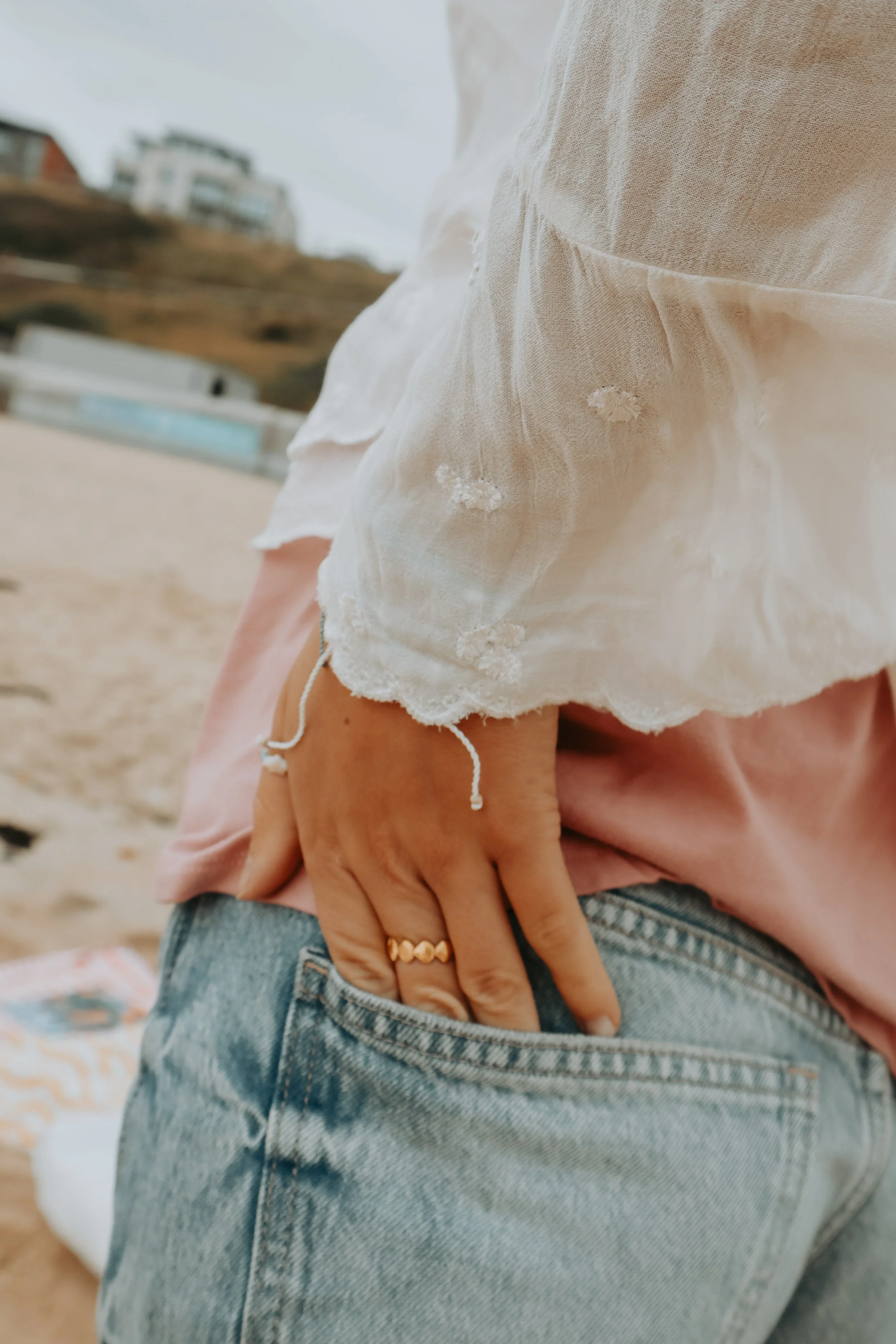 Close-up of a person wearing a white, embroidered, semi-sheer blouse and ripped denim jeans. The person's hand, adorned with rings and a bracelet, is resting on their hip, with a beach and buildings visible in the blurred background.