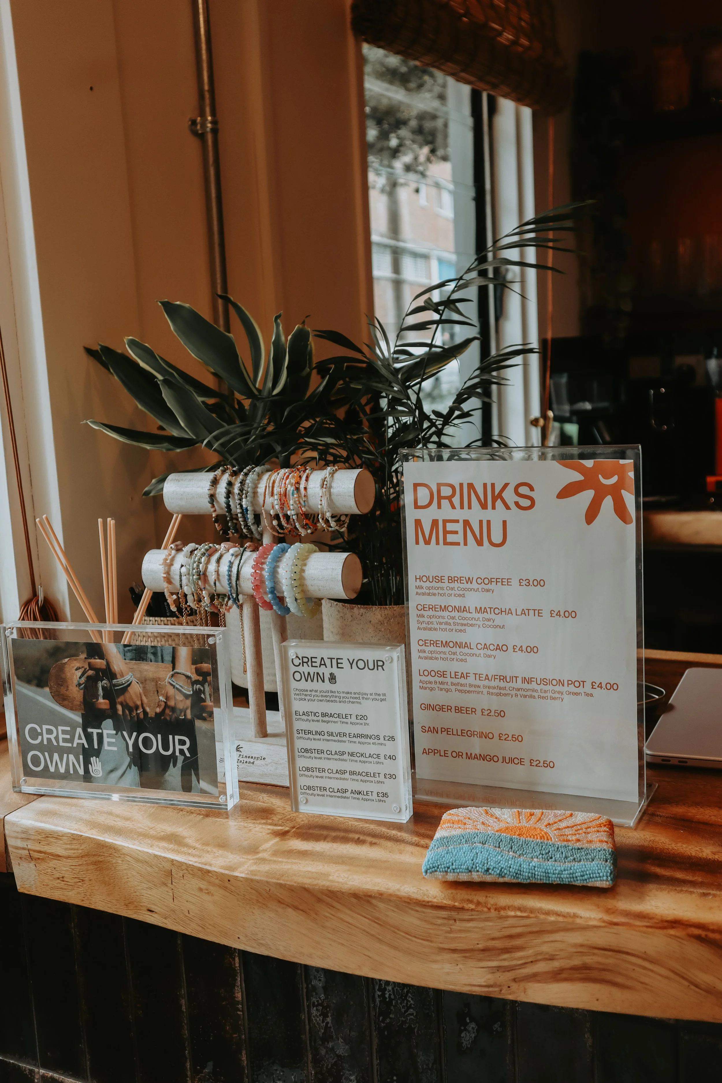 A wooden counter with jewelry display, a plant, and a drinks menu listing beverages like house brew coffee, matcha latte, and various infusions, along with prices. Pineapple Island Jewellery Photography