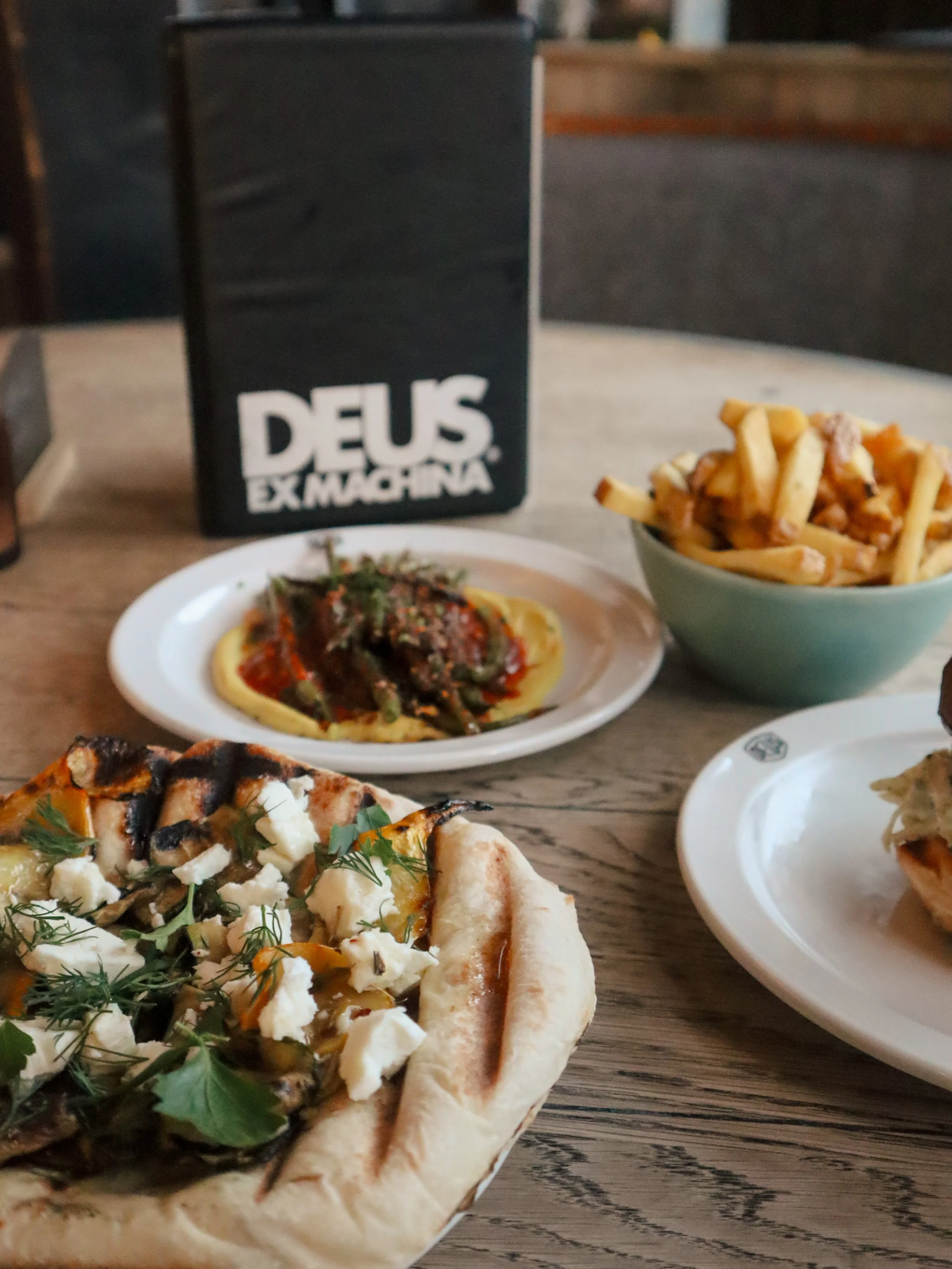 A table with various dishes including a pizza, a bowl of fries, and a small bowl of pasta, with a black drink container labeled "Deus Ex Machina" in the background.