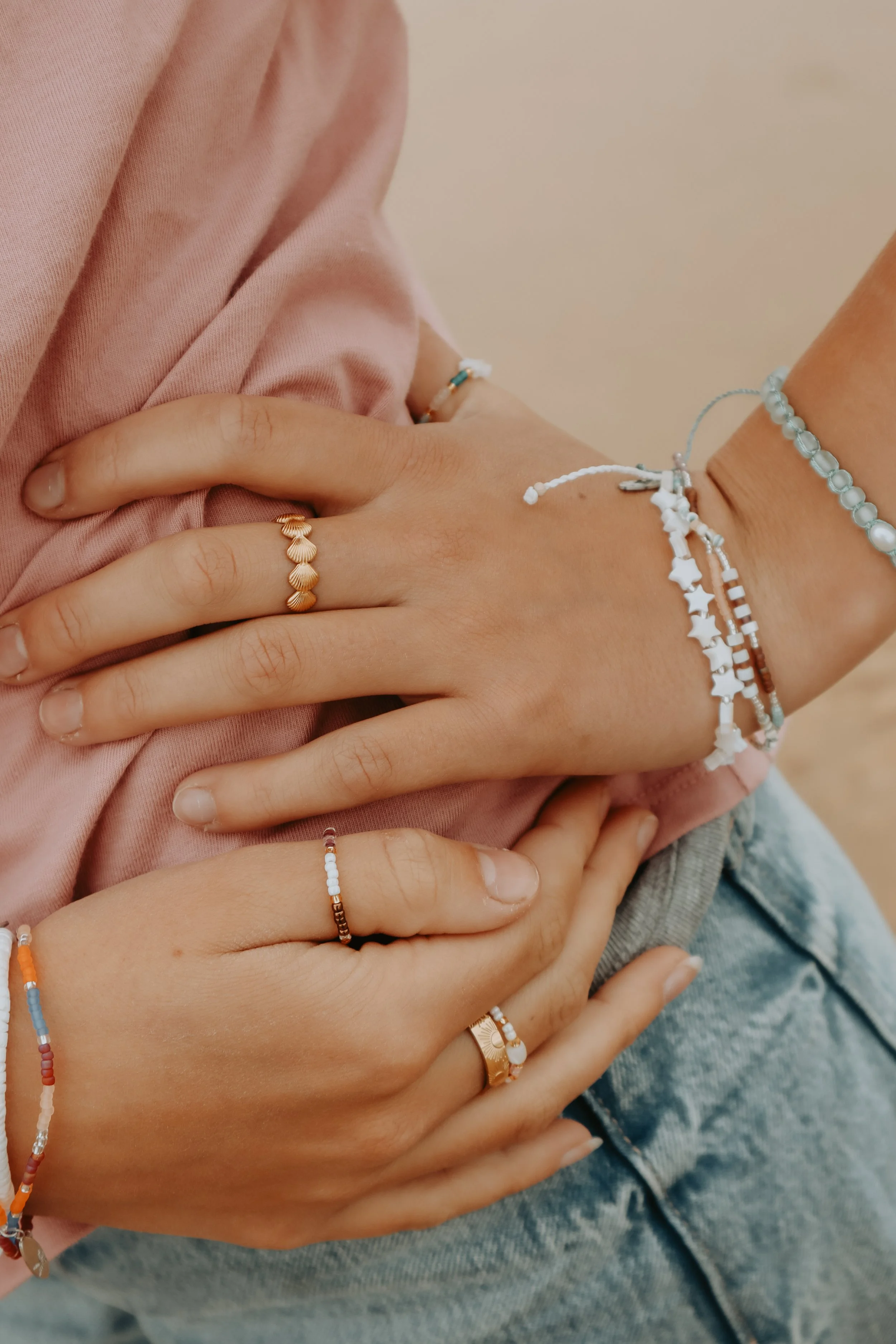 Close-up of multiple hands with various rings and bracelets, resting on a person's lap wearing a pink shirt and jeans.