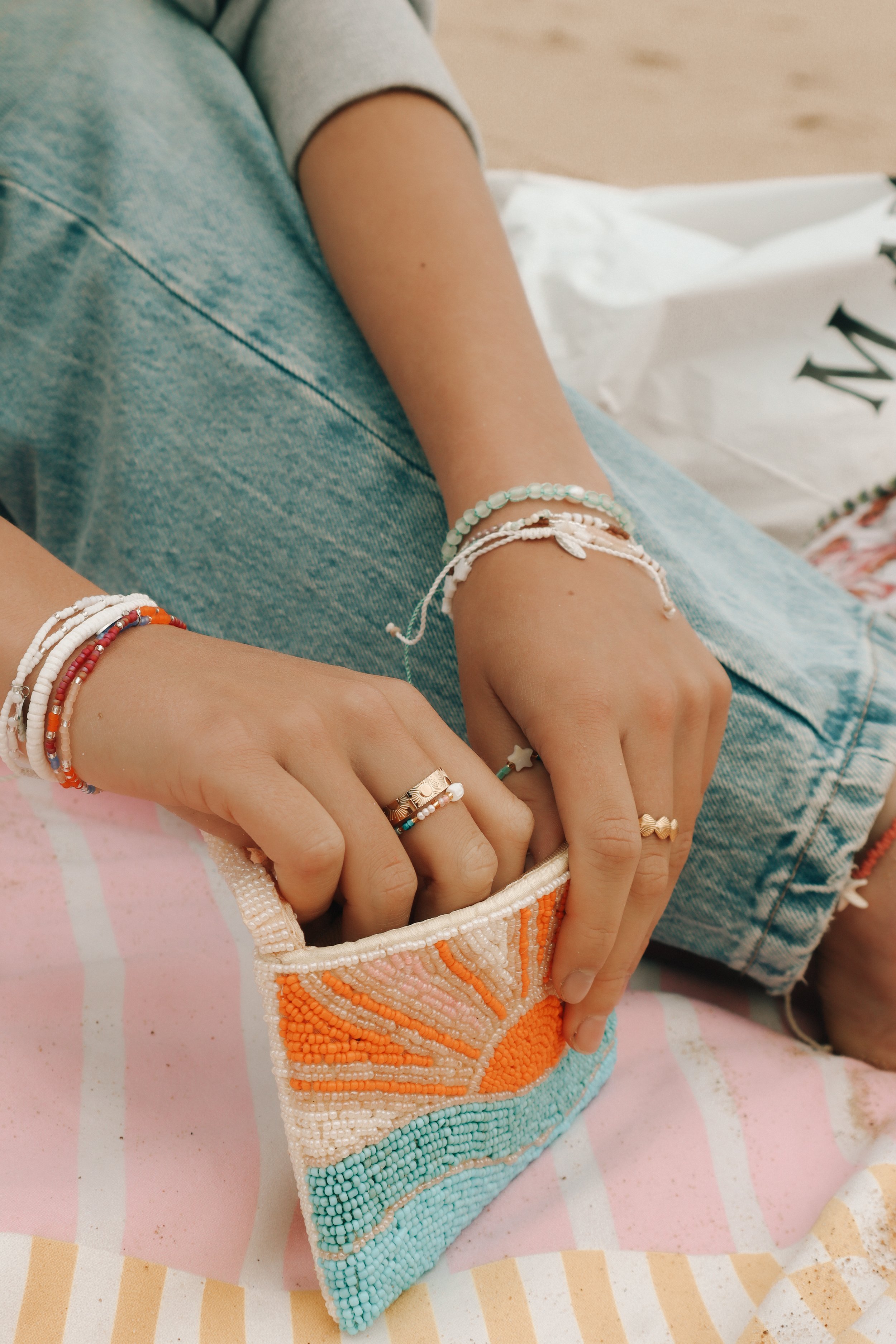 Person wearing multiple beaded bracelets, sitting on a striped pink and white towel, with a colorful beaded clutch in their hands, and a shopping bag nearby.