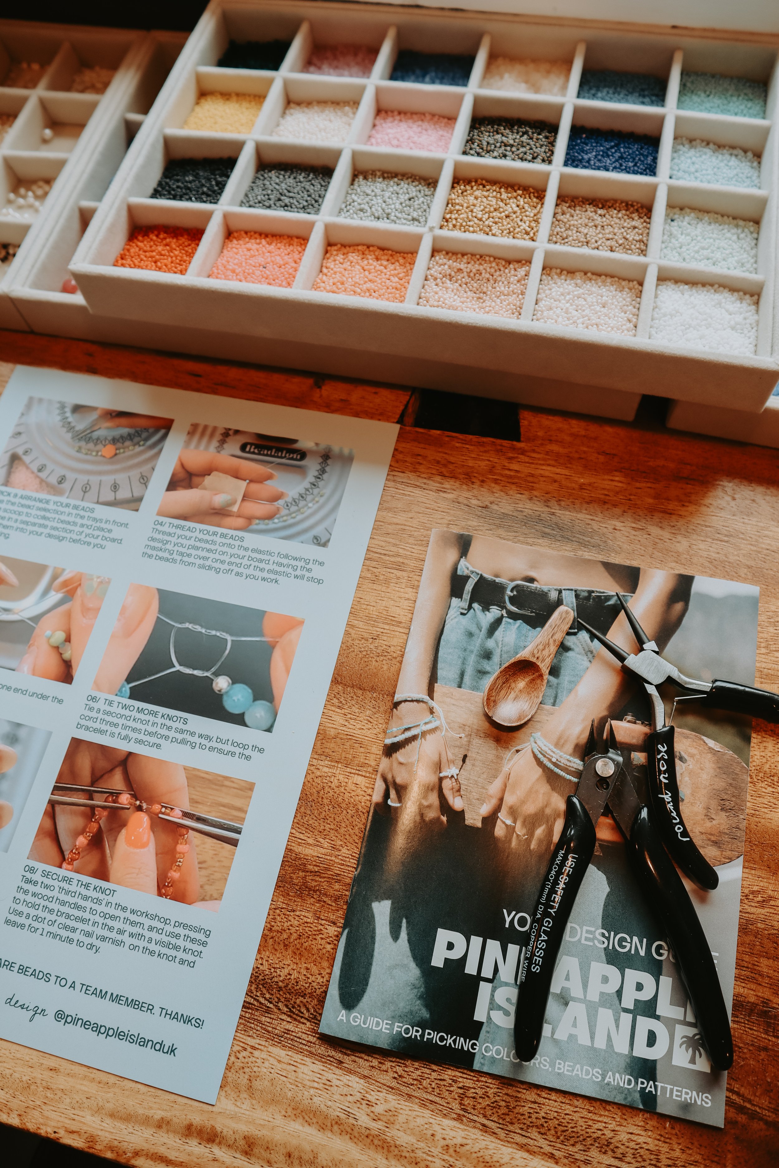 A wooden jewelry making kit with small compartments filled with multicolored beads. On the table, there are instruction sheets with images of jewelry and a pamphlet titled 'Pineapple Island' about picking colors, beads, and patterns for jewelry makin