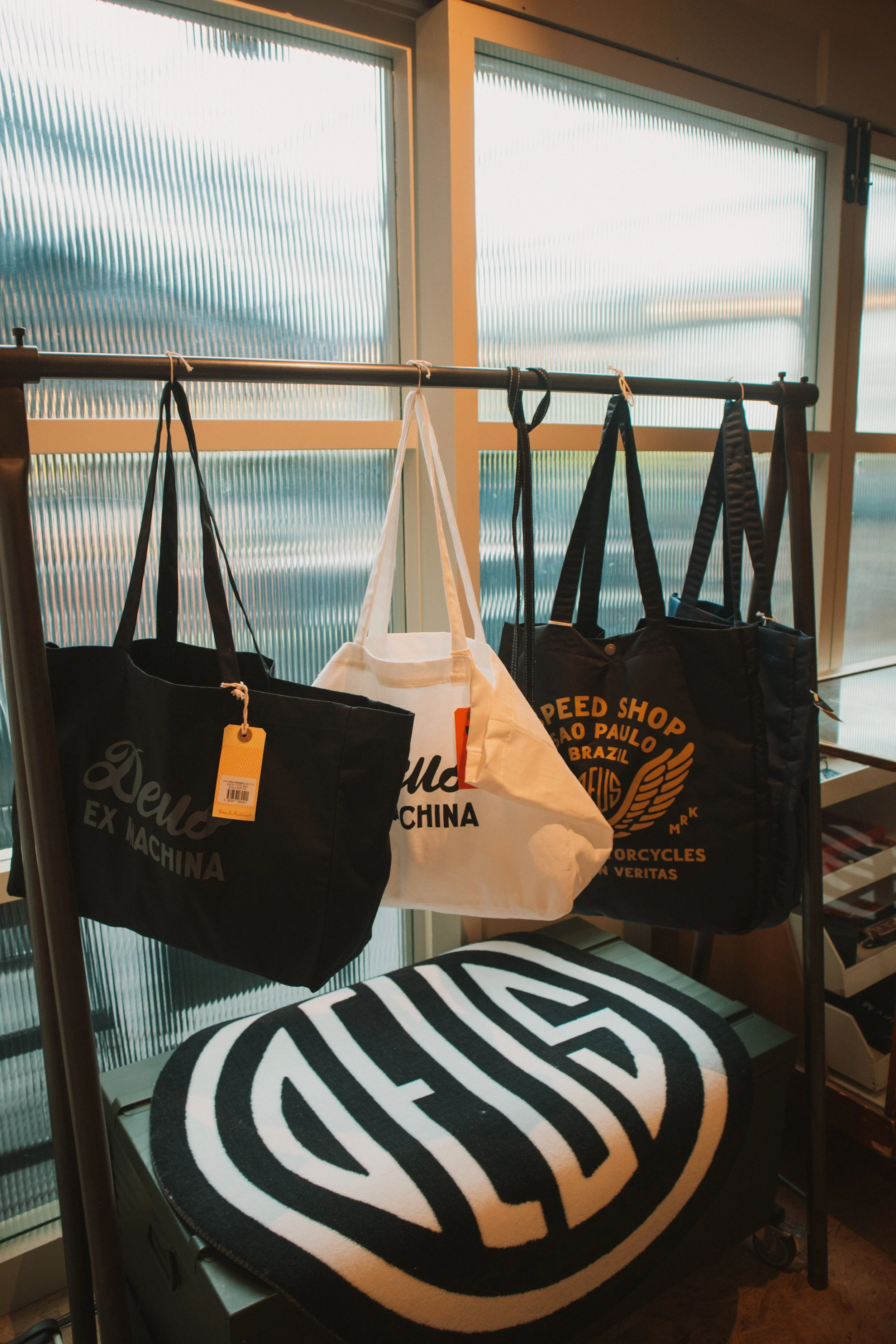 A display rack with four tote bags hanging on hooks in front of large frosted glass windows. The tote bags are black, white, and black with orange and yellow text. The black tote bags have varying text, some reading 'Deus Ex Machina,' and one has a y
