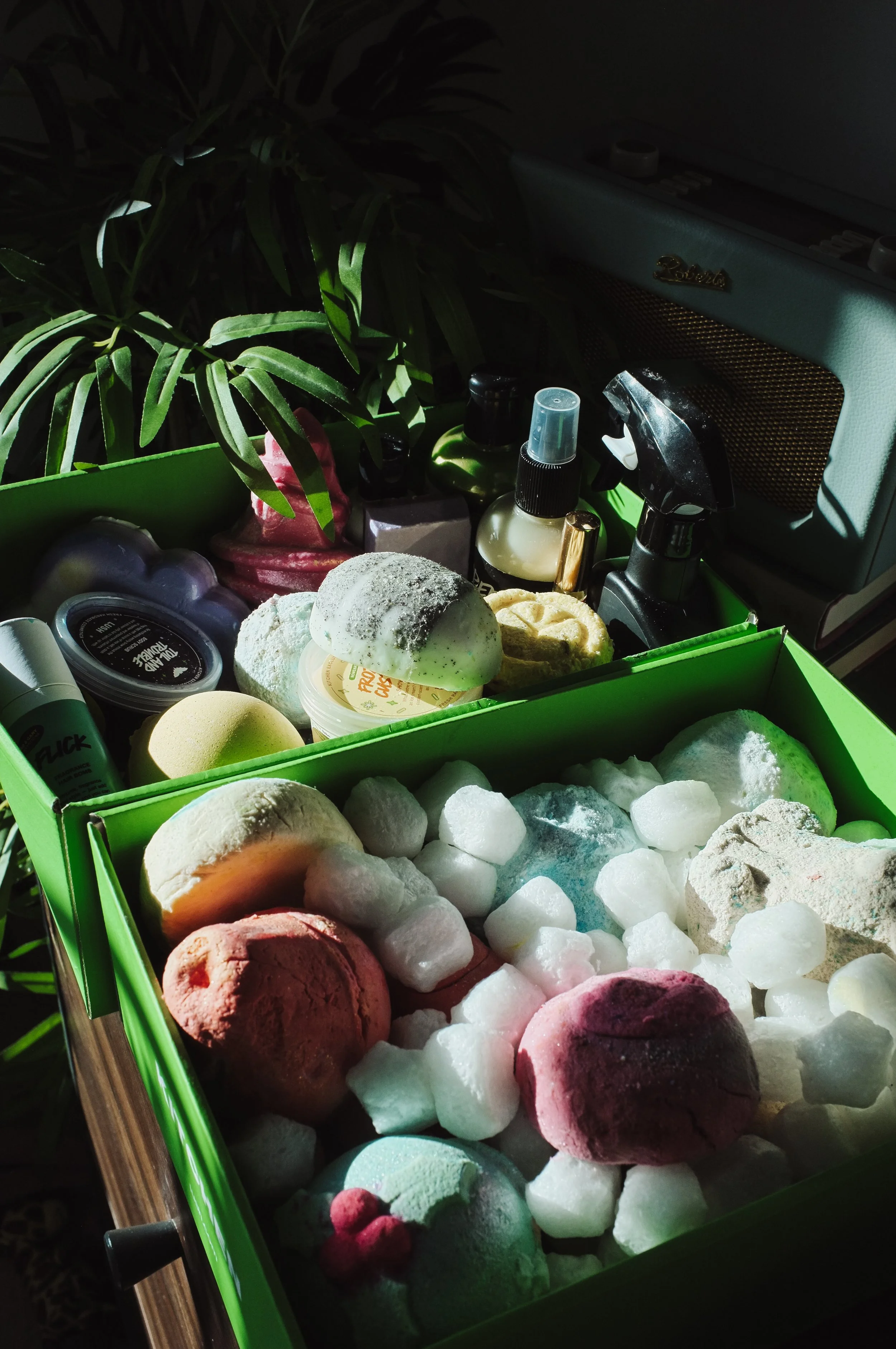 A green box filled with various colorful bath bombs and soap bars on ice, with surrounding skincare bottles and an old-fashioned radio in the background.