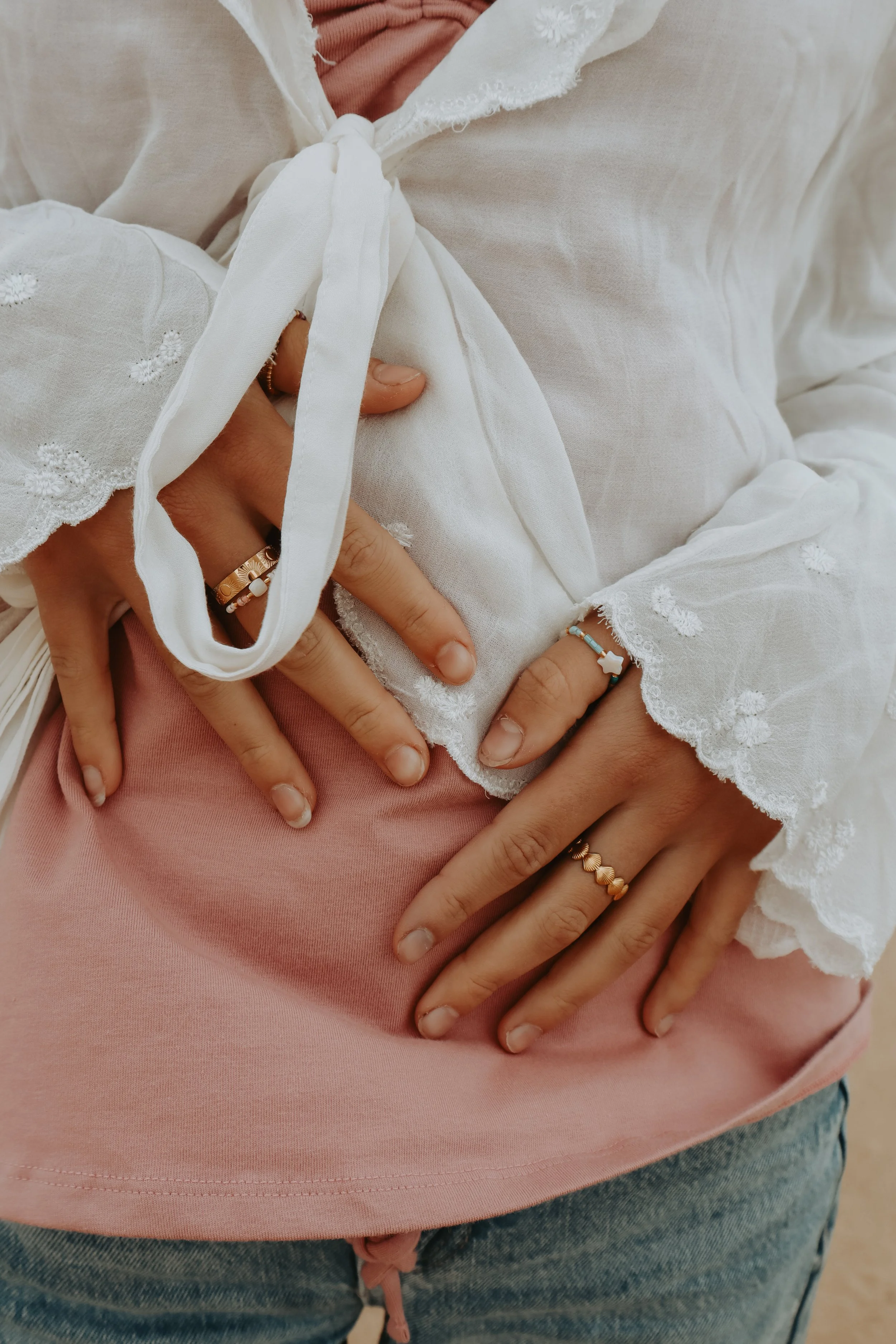 Close-up of hands intertwined, showing rings and bracelets, with part of a white embroidered blouse and pink shirt.