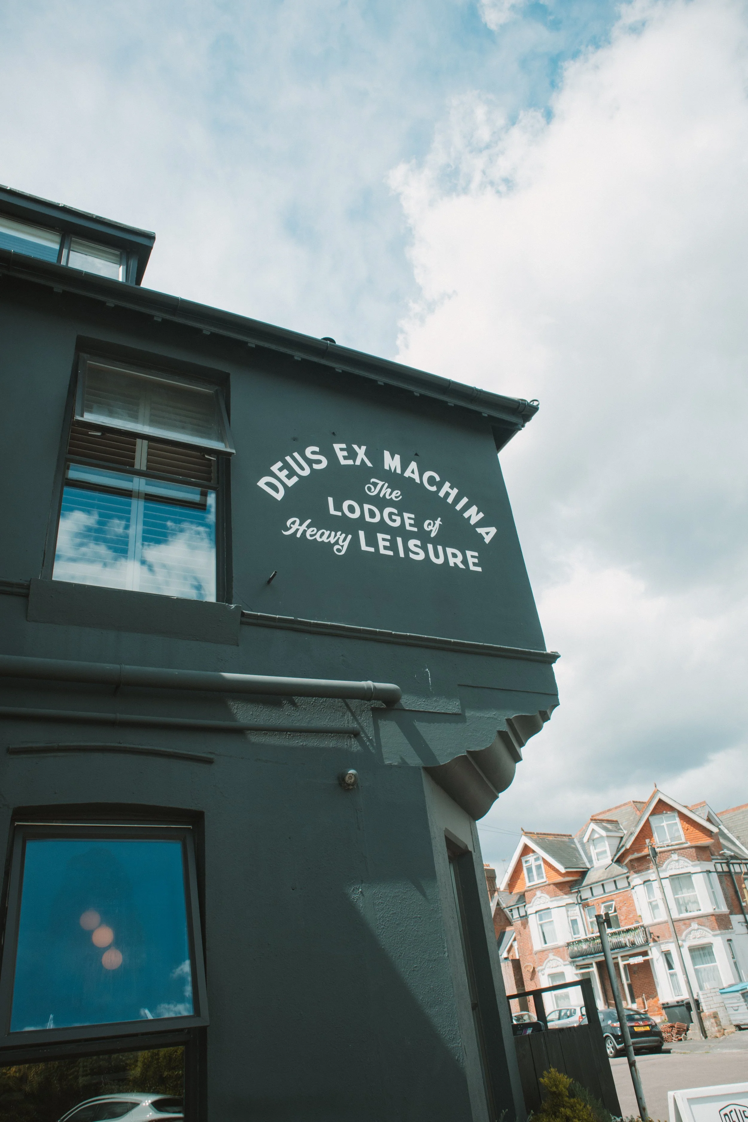 A gray building with a sign that reads 'Deus Ex Machina, The Lodge of Heavy Leisure,' with windows reflecting the cloudy sky. Residential buildings with orange rooftops are visible in the background.
