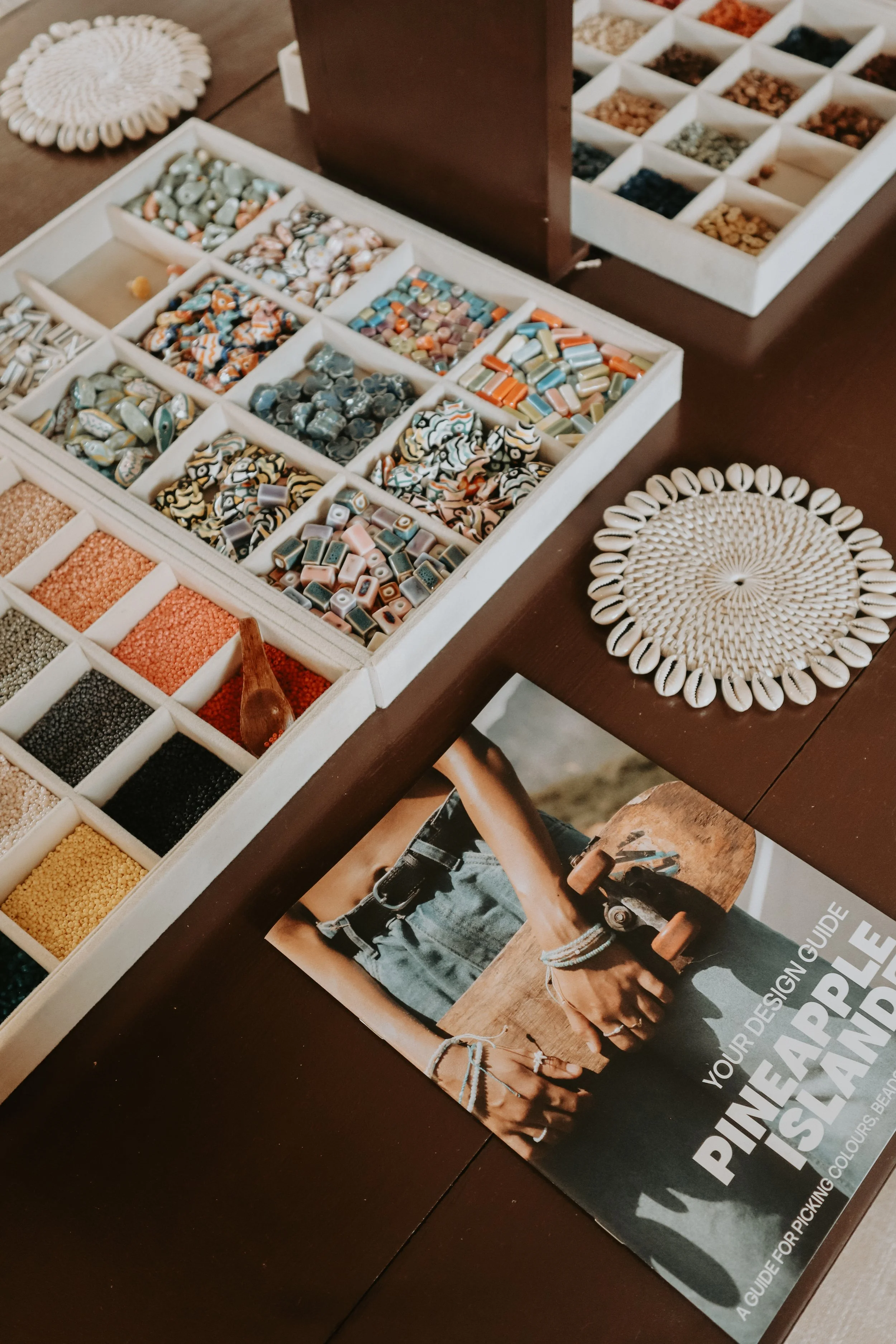 A display of colorful beads in a divided white tray, with a selection of seashells arranged in a circle on a dark wooden table. A fashion magazine featuring a person holding a skateboard is partially visible underneath the tray.