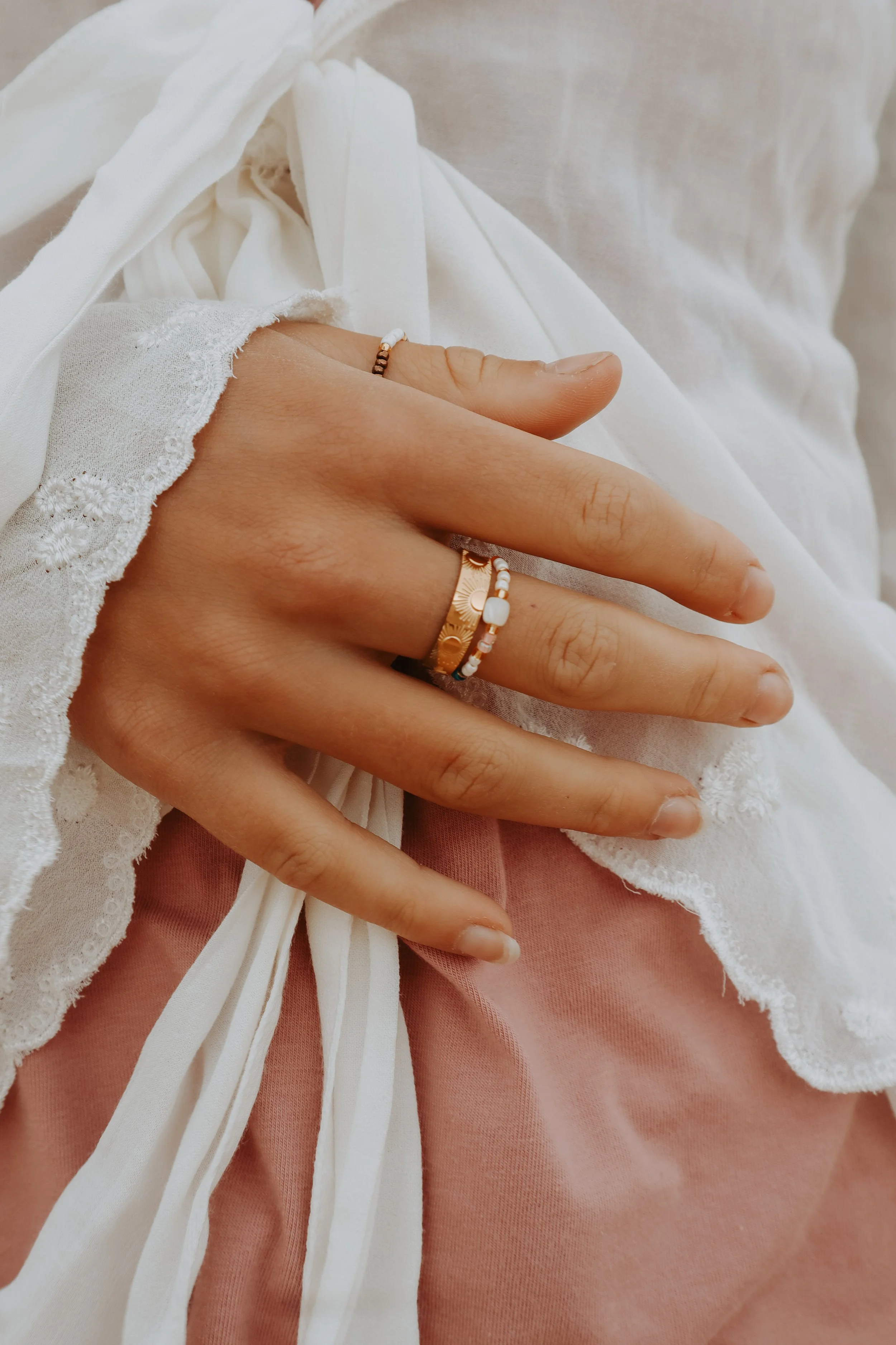 A person's hand wearing rings resting on their lap, with white lace-trimmed fabric and pink clothing visible.