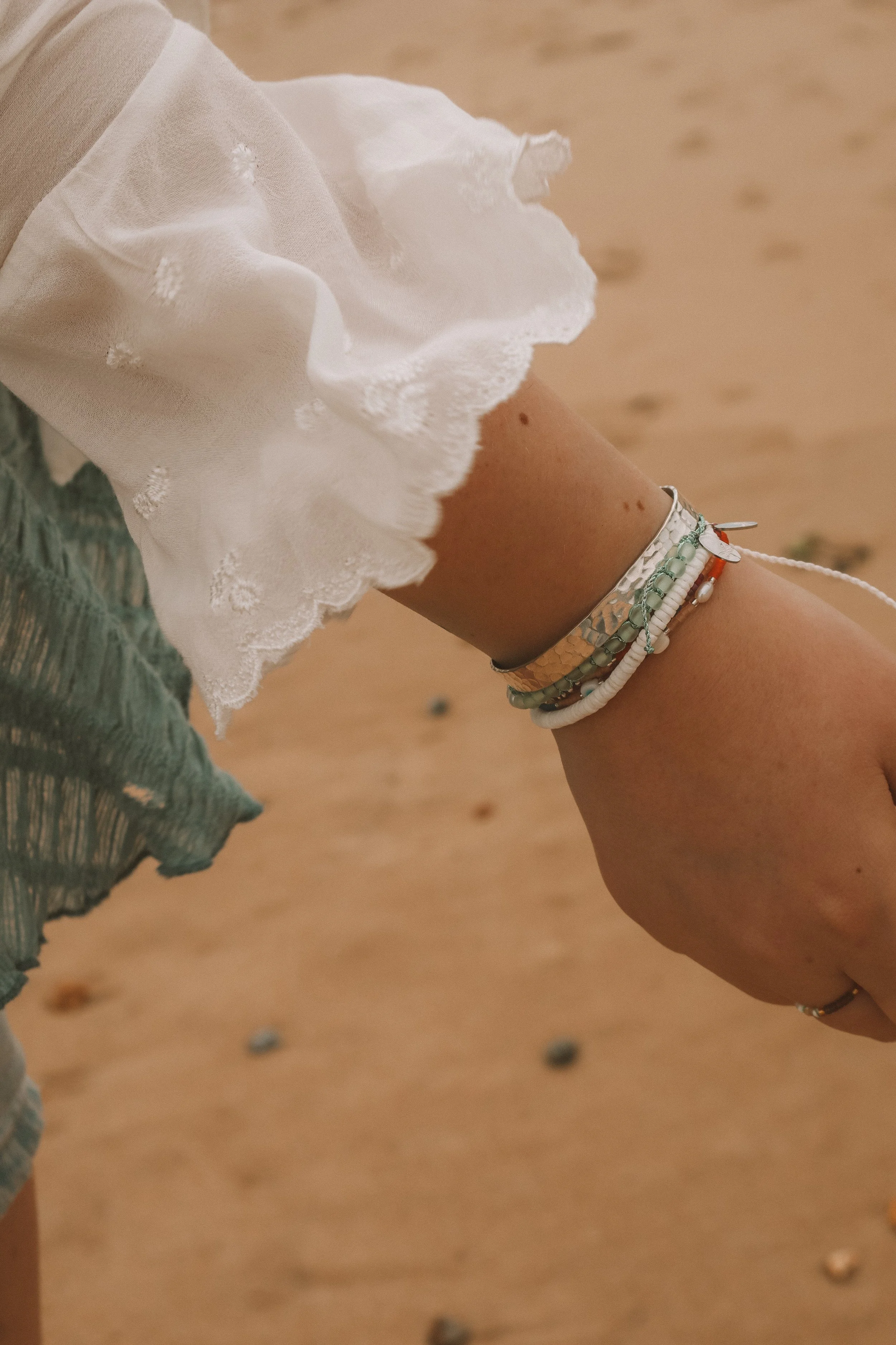 Close-up of a person wearing multiple colorful beaded bracelets on their wrist, with a white, frayed fabric top and a sandy beach background.
