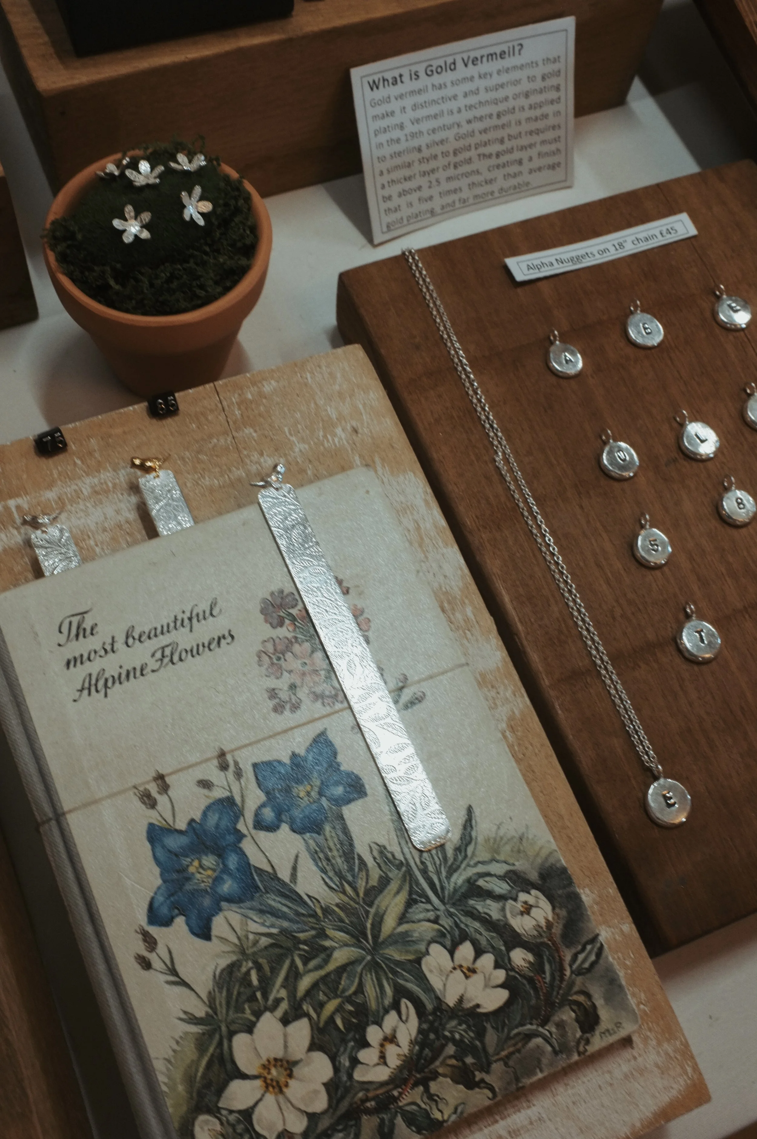 Display of silver jewelry, including pendants, necklaces, and charms, with informational signs about gold vermeil and alpine flowers, alongside a potted plant with silver flower earrings.