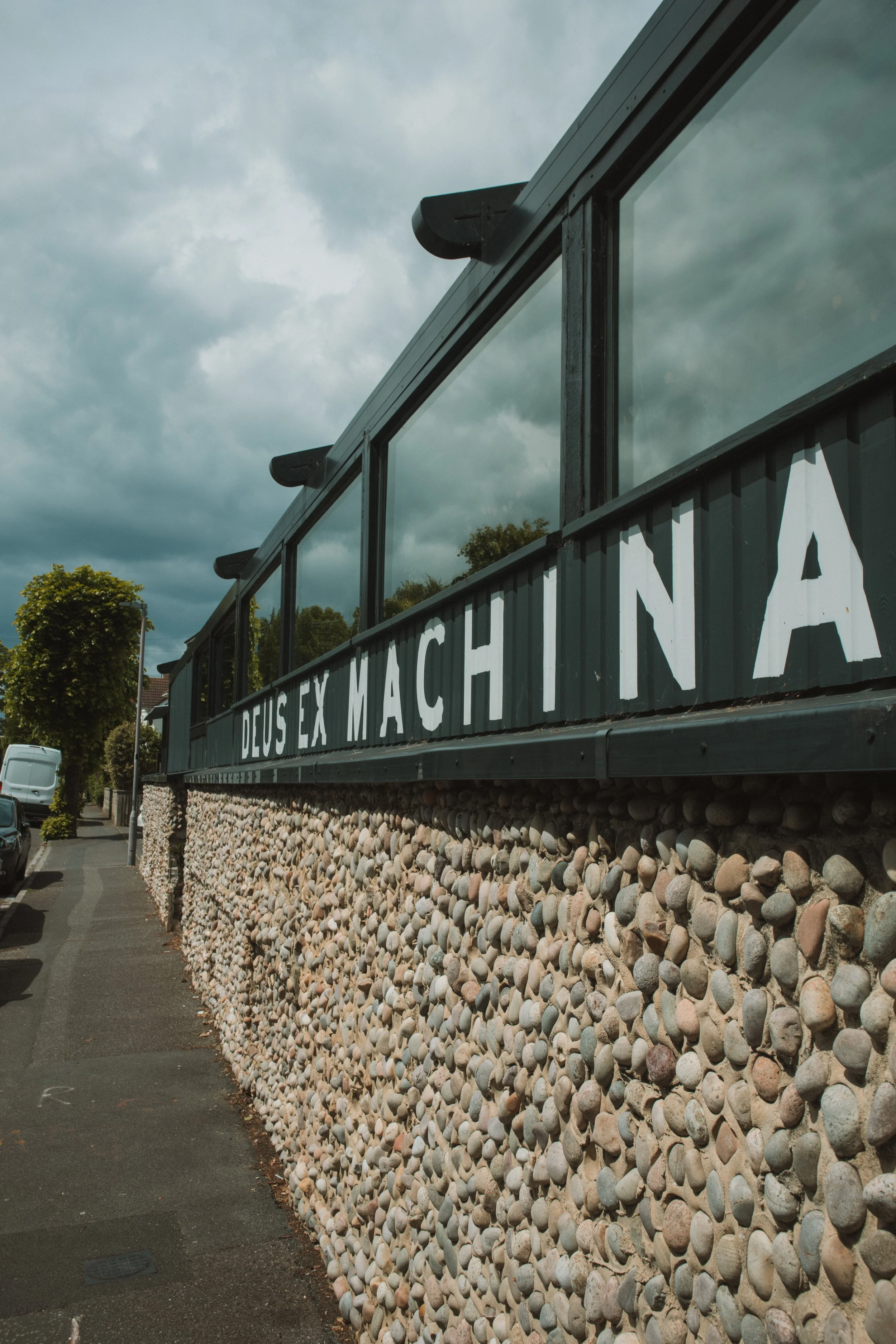 A building with a sign that reads 'Deus Ex Machina' on a stone wall beneath a cloudy sky.