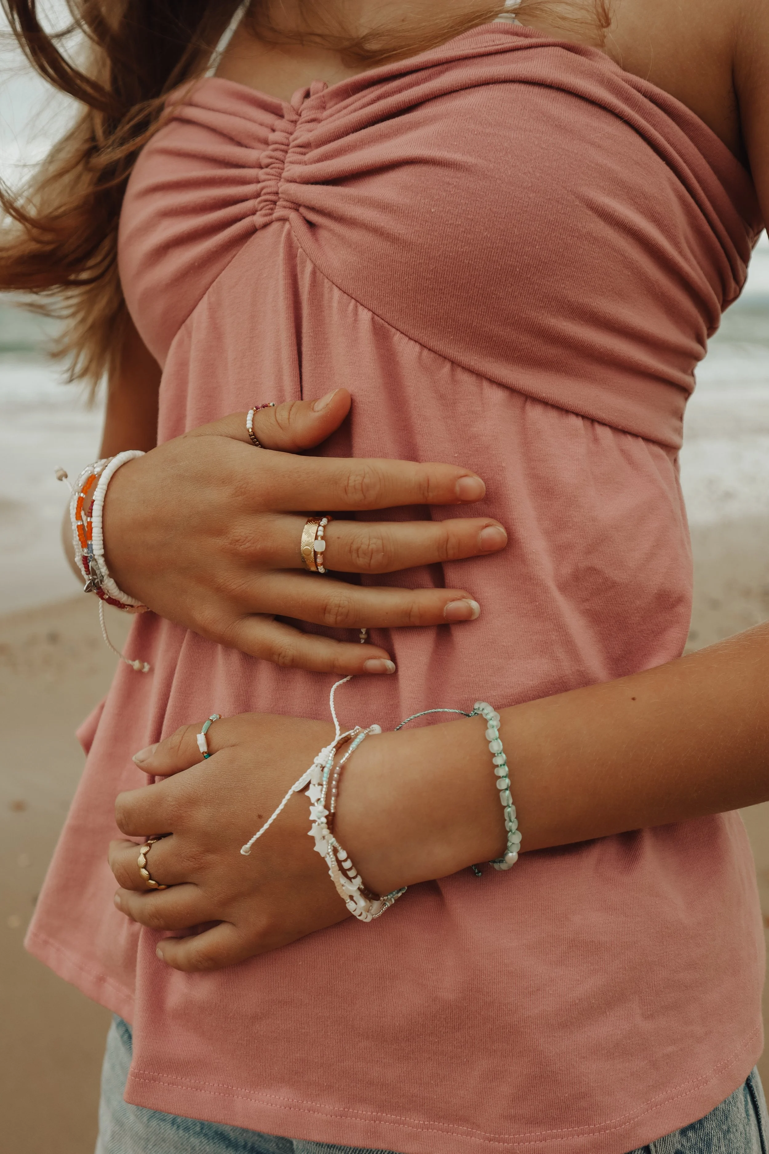 Close-up of a person wearing a pink top with ruched details on the shoulder, holding their waist with both hands. The person is wearing multiple beaded bracelets and rings, and has wavy hair. The background shows a beach setting with sand and ocean w