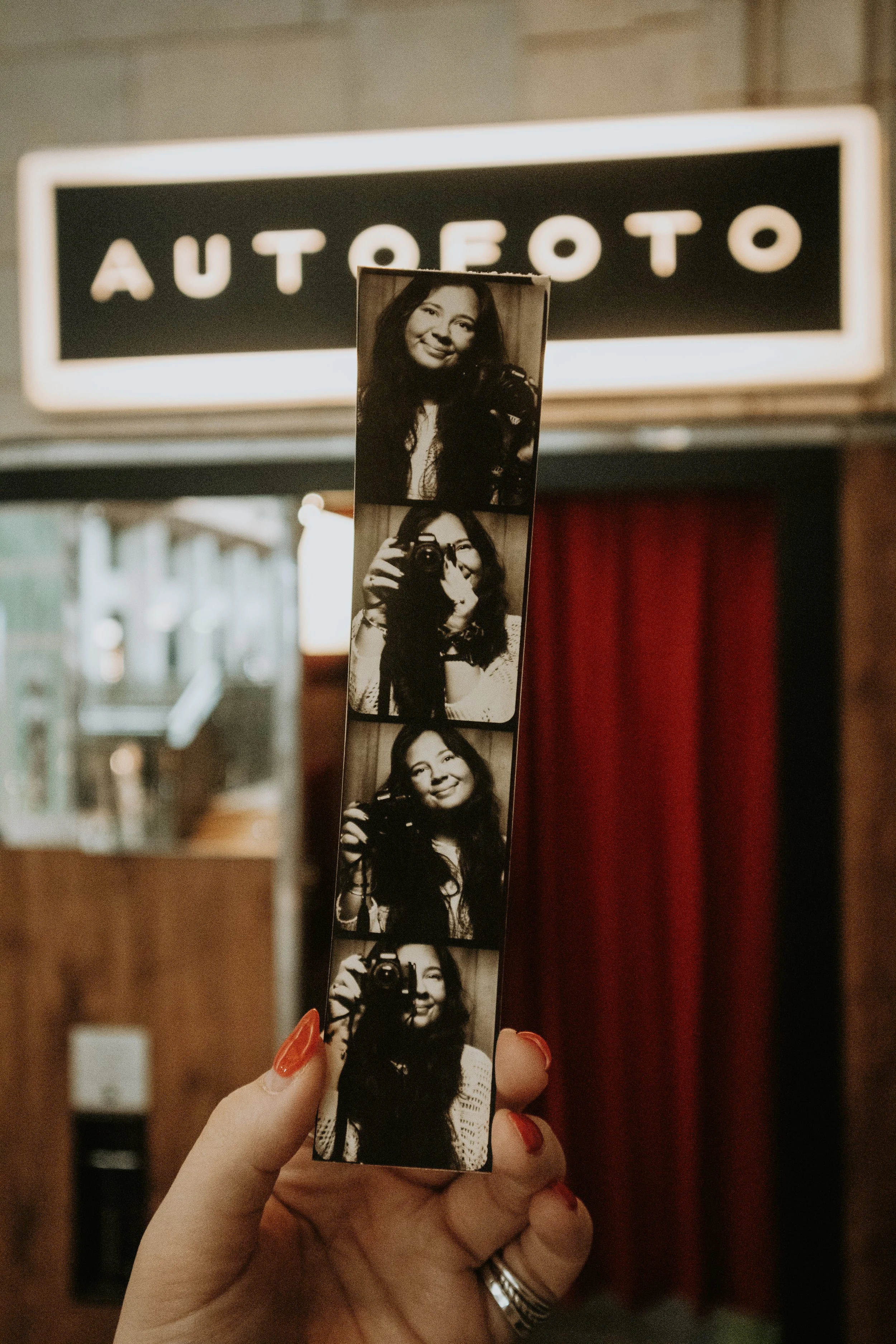 A person with red nail polish holding a strip of black and white photographs of a woman with long hair and a camera, in front of a sign that reads 'AUTOEOTO'.