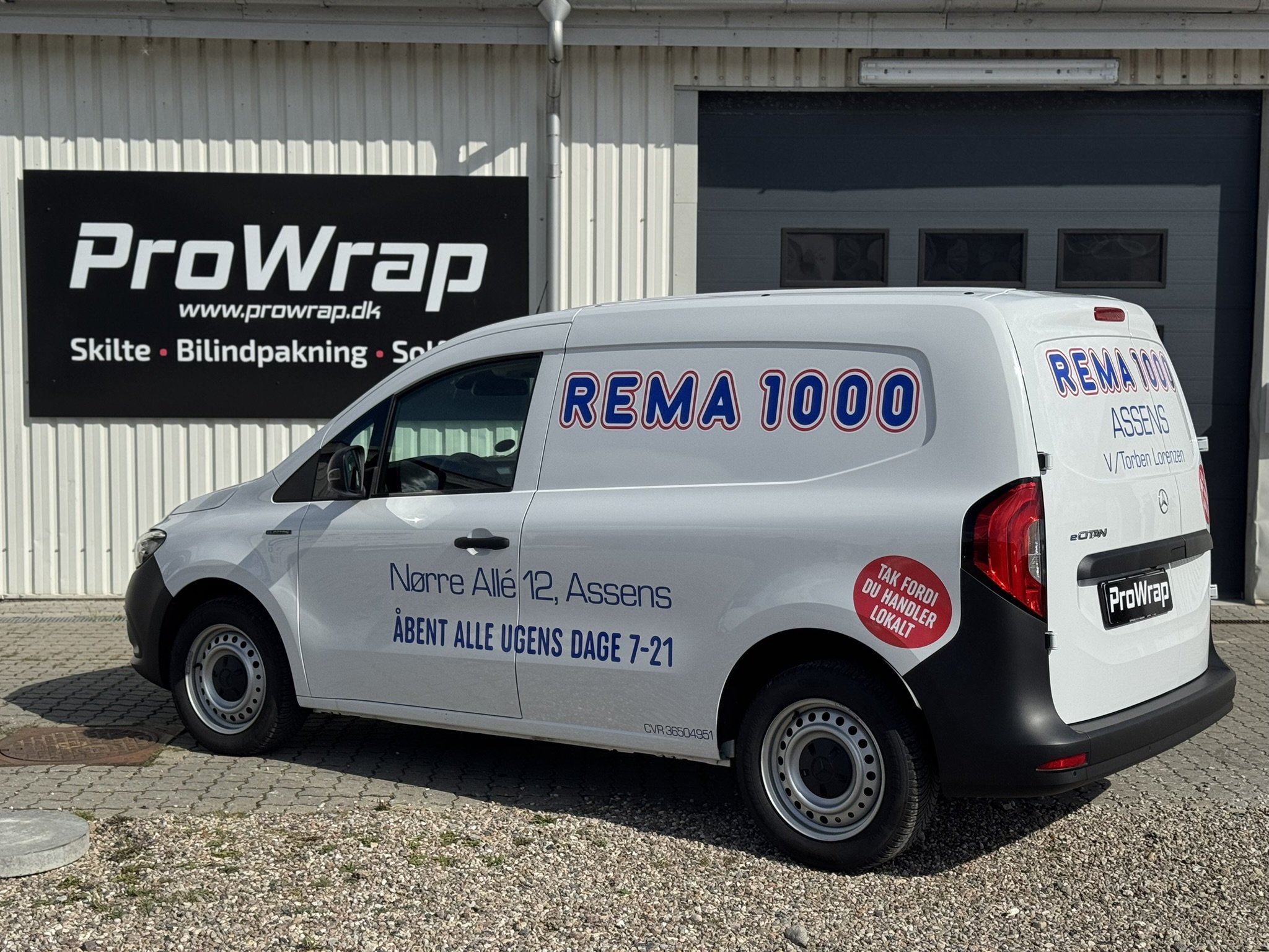 En hvid firmabil med teksten 'REMA 1000' og adresse 'Nørre Allé 12, Assens' står foran en stor lagerbygning med skiltet 'ProWrap' og tekst om skilte, bilindpakning og solfilm.