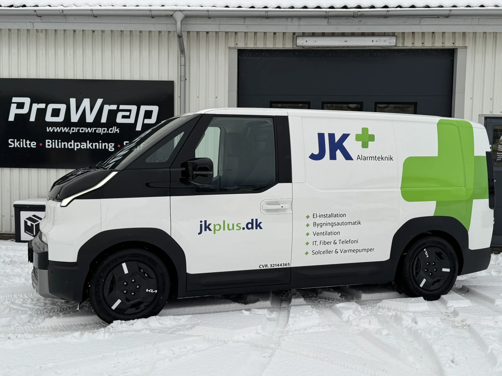 Hvid og sort firmabil med grøn detaljer, der reklamerer for JK Alarmteknik, parkeret foran en adresse med en sort facadeskilt forprowap.dk.