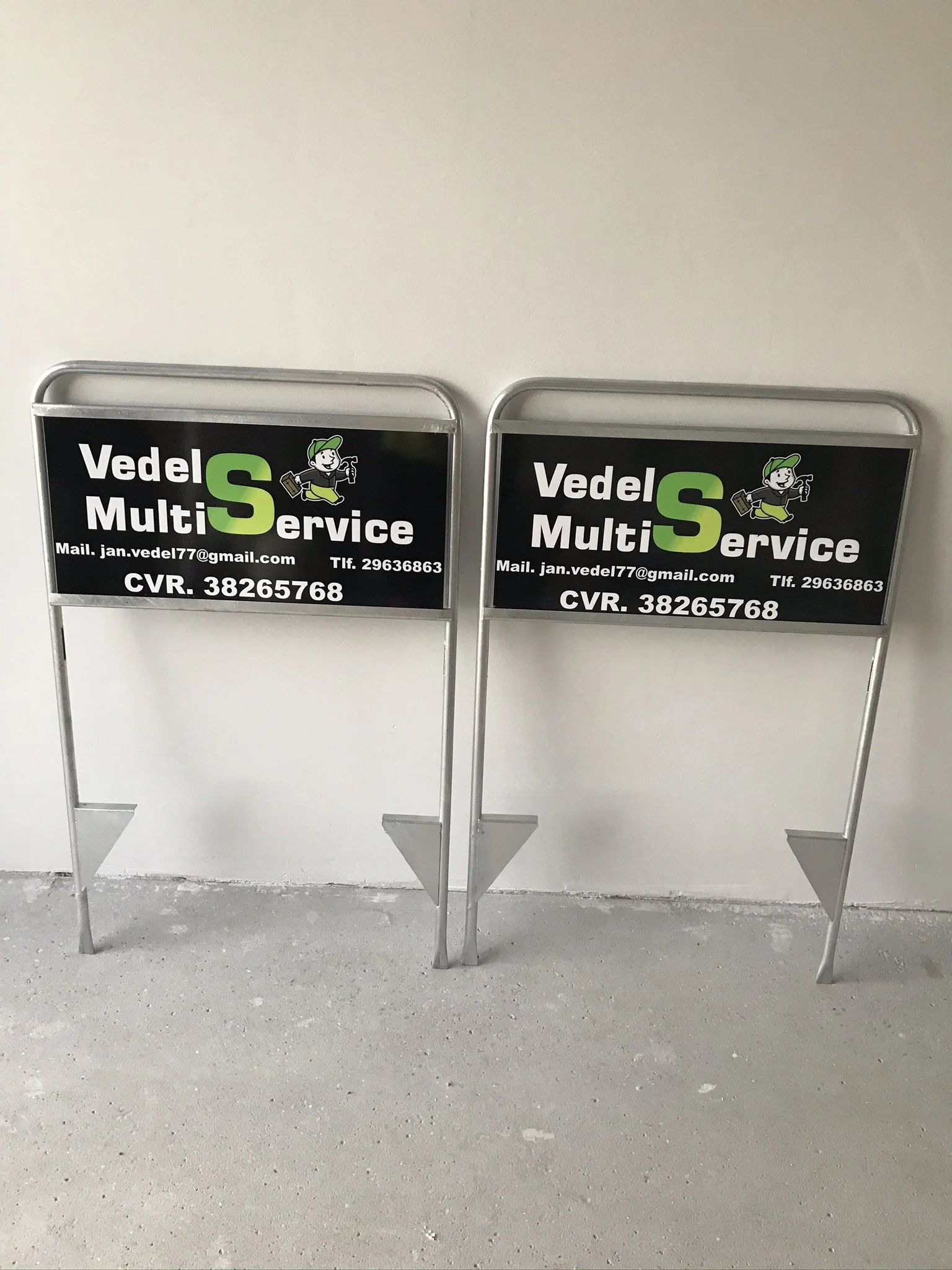 To metal reklameskilte med logo og kontaktinformationer for Vedel Multi Service, fastgjort til en metalstød på en hvid væg.
