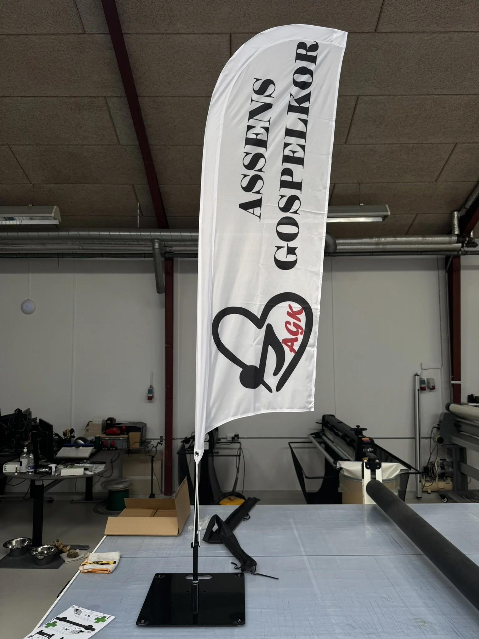 En sort base med en justerbar arm som holder en hvid flag med sort tekst og logo. Baggrunden er en industrielt indrettet værksted med borde, værktøj og udstyr.