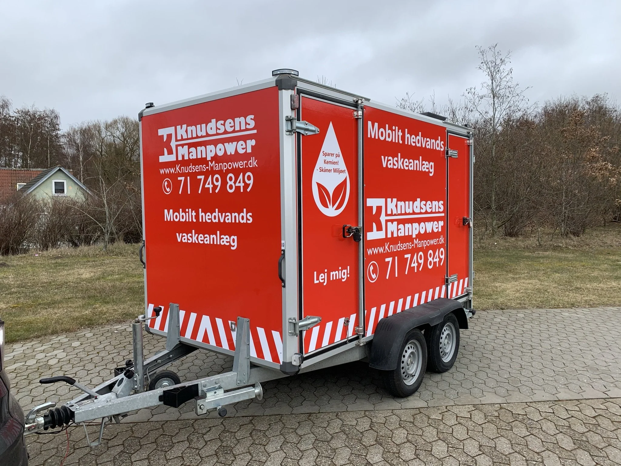 Rød mobil vaskeanlægs- og kemikalieservice trailer med tekst og logo fra Knudsens Manpower, parkeret på en brostensbelagt vej med et hus og træer i baggrunden.
