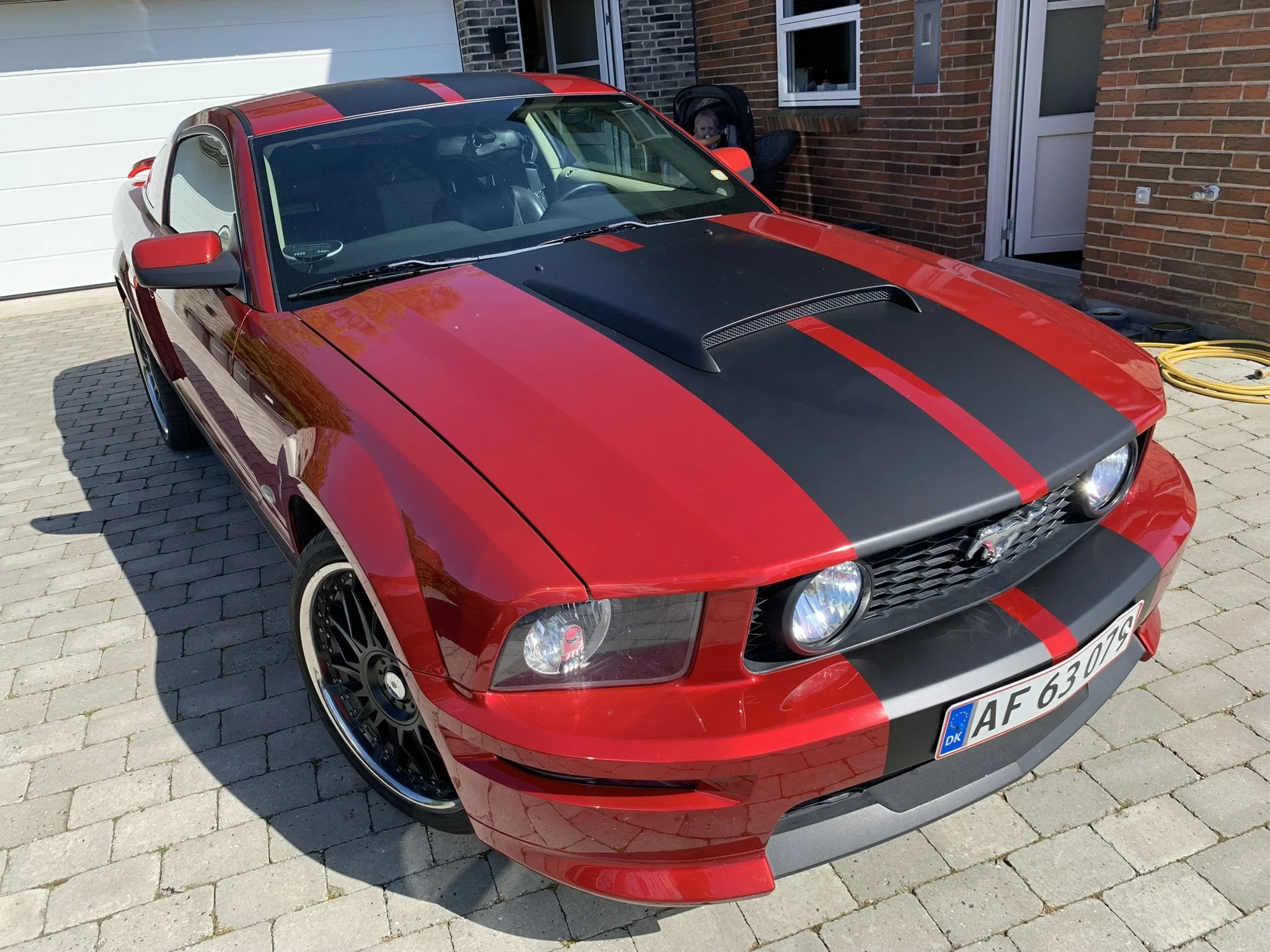 En rød sportsvogn med sort hætte og to brede sorte striber på taget er parkeret foran en bolig med murstensfacade. En baby sidder i en barnevogn i baggrunden nær en dør.