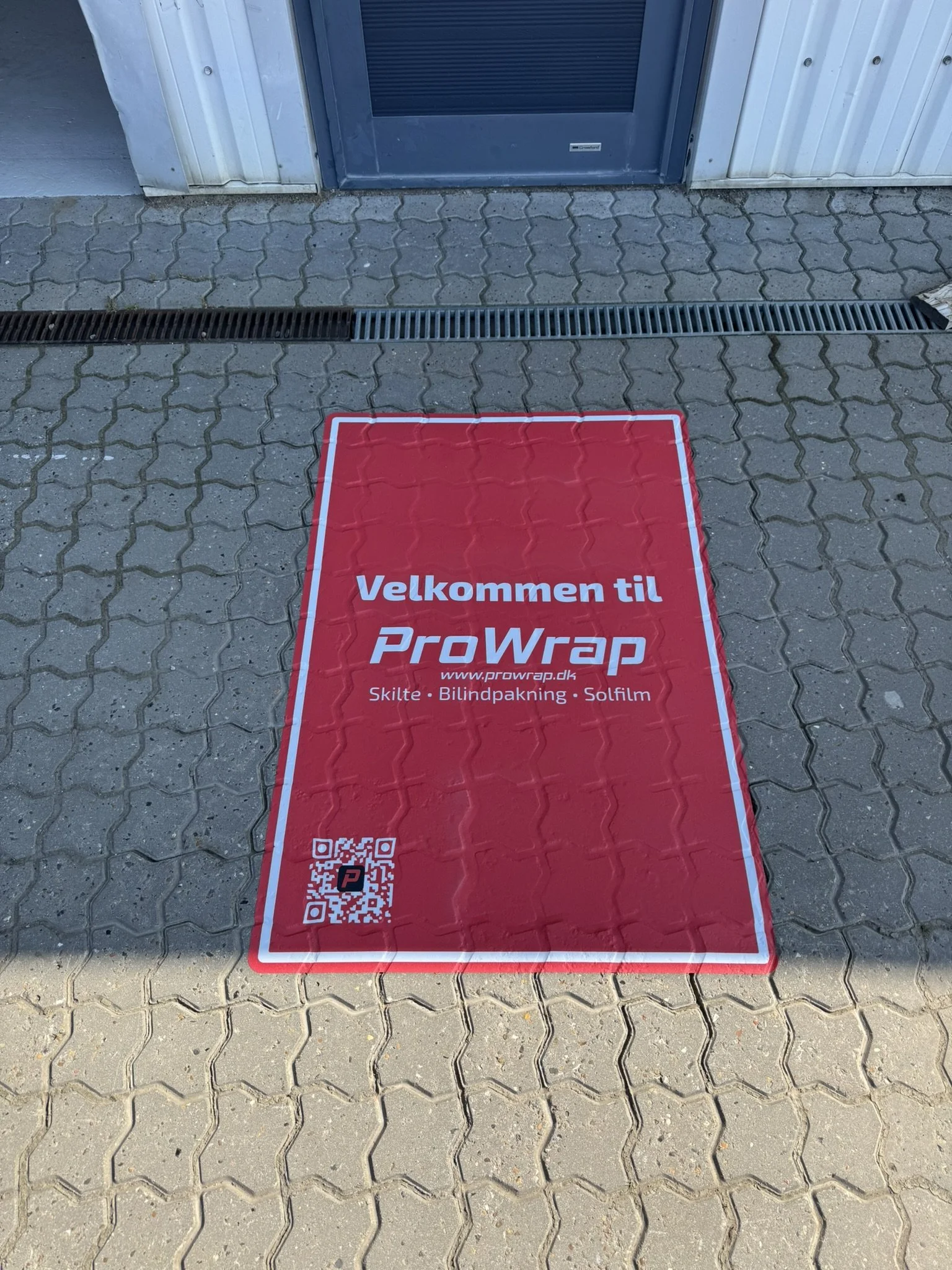 Et rødt tæppe med teksten 'Velkommen til ProWrap' ligger på fortovet foran en bygning med en sort dør og en metalklap til afløb.
