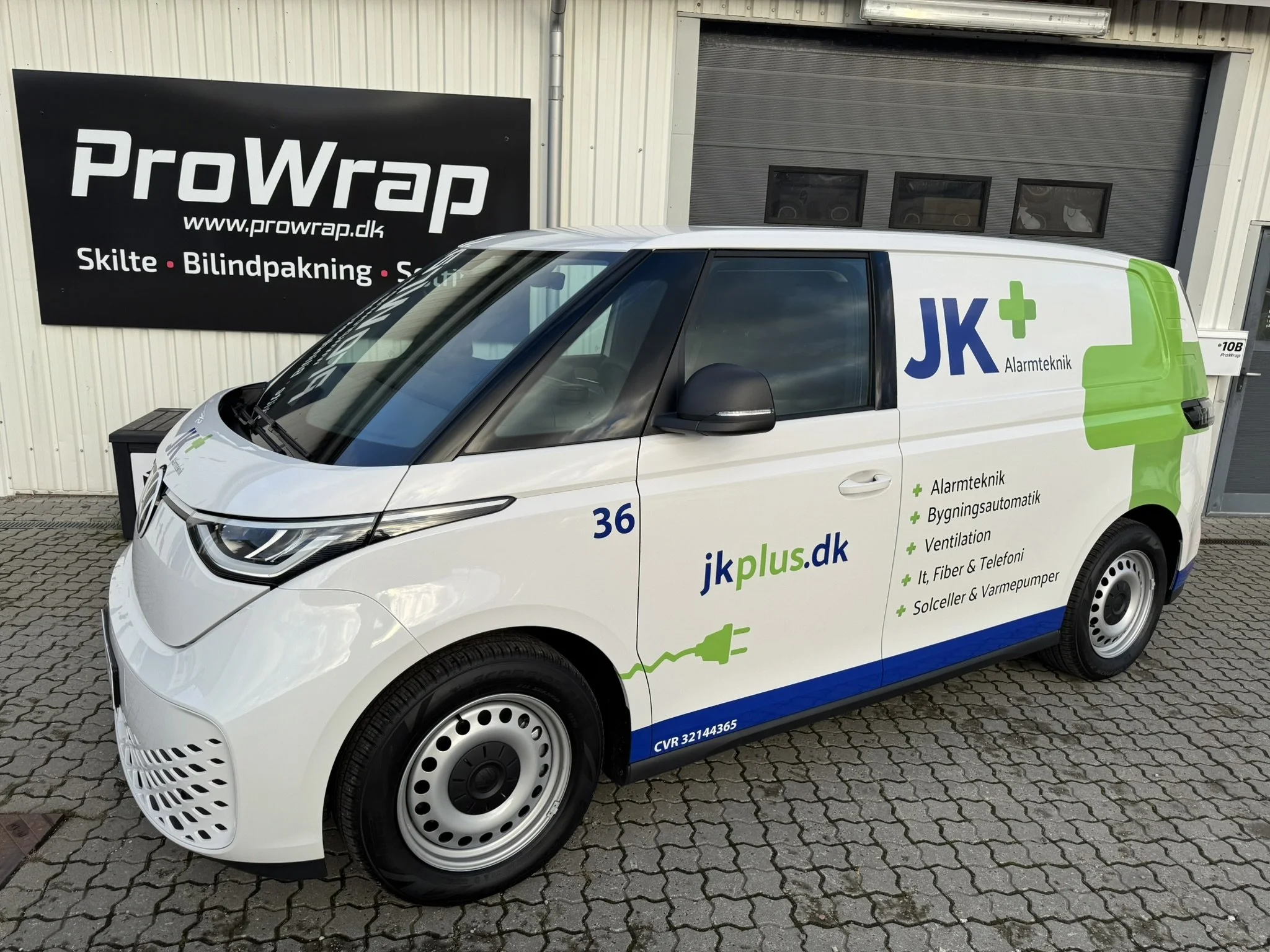 En hvid varebil med grøn og blå logoer, der reklamerer for JK Alarmteknik, parkeret foran en bygning med reklameskilt for ProWrap, en skilteteknikvirksomhed.
