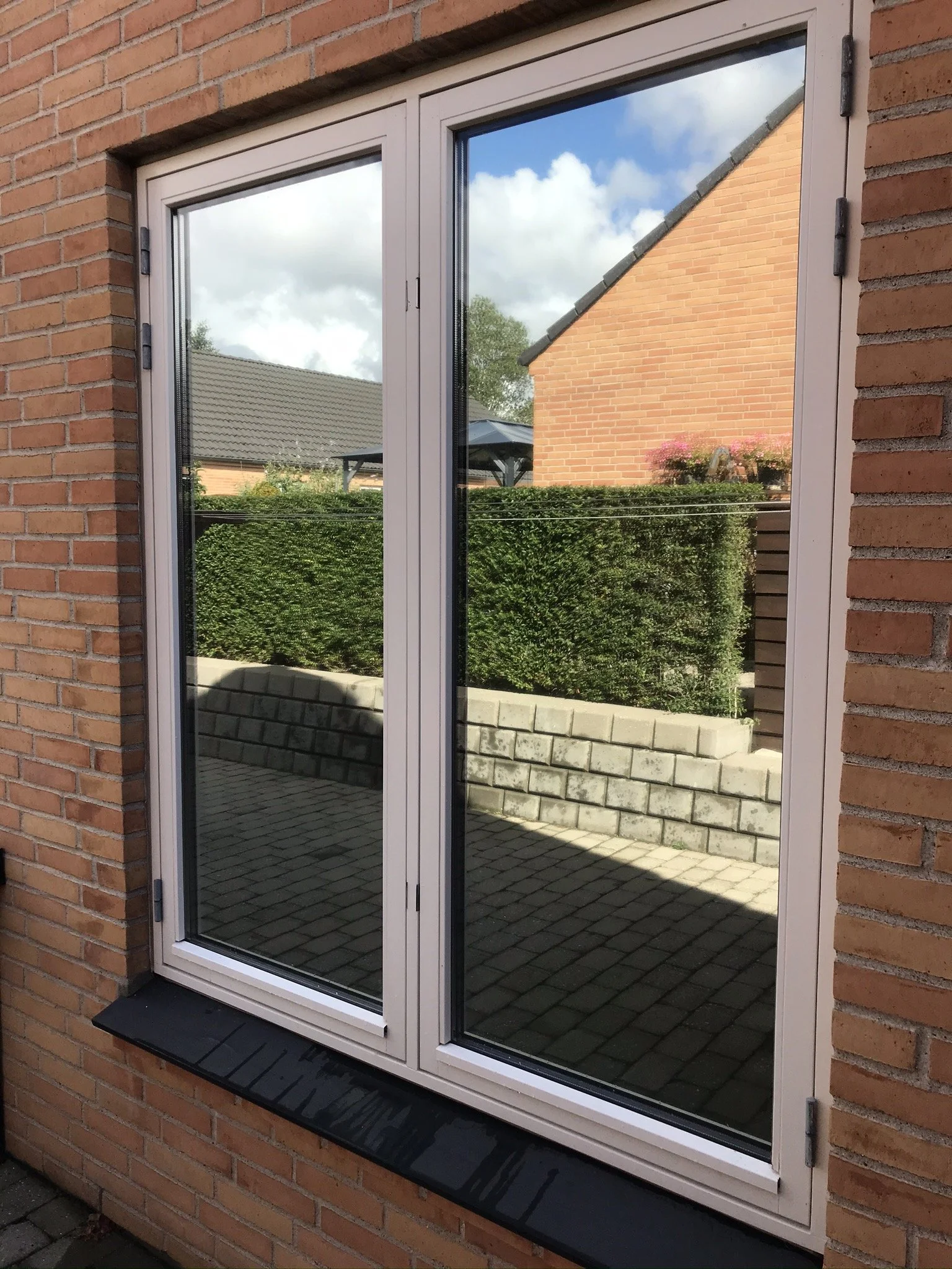 Dobbeltvindue i rød murstensvæg, med reflekterende glas, der viser en baghave med græs, buske og en tatovering. Under vinduet er en sort skiferbrik.
