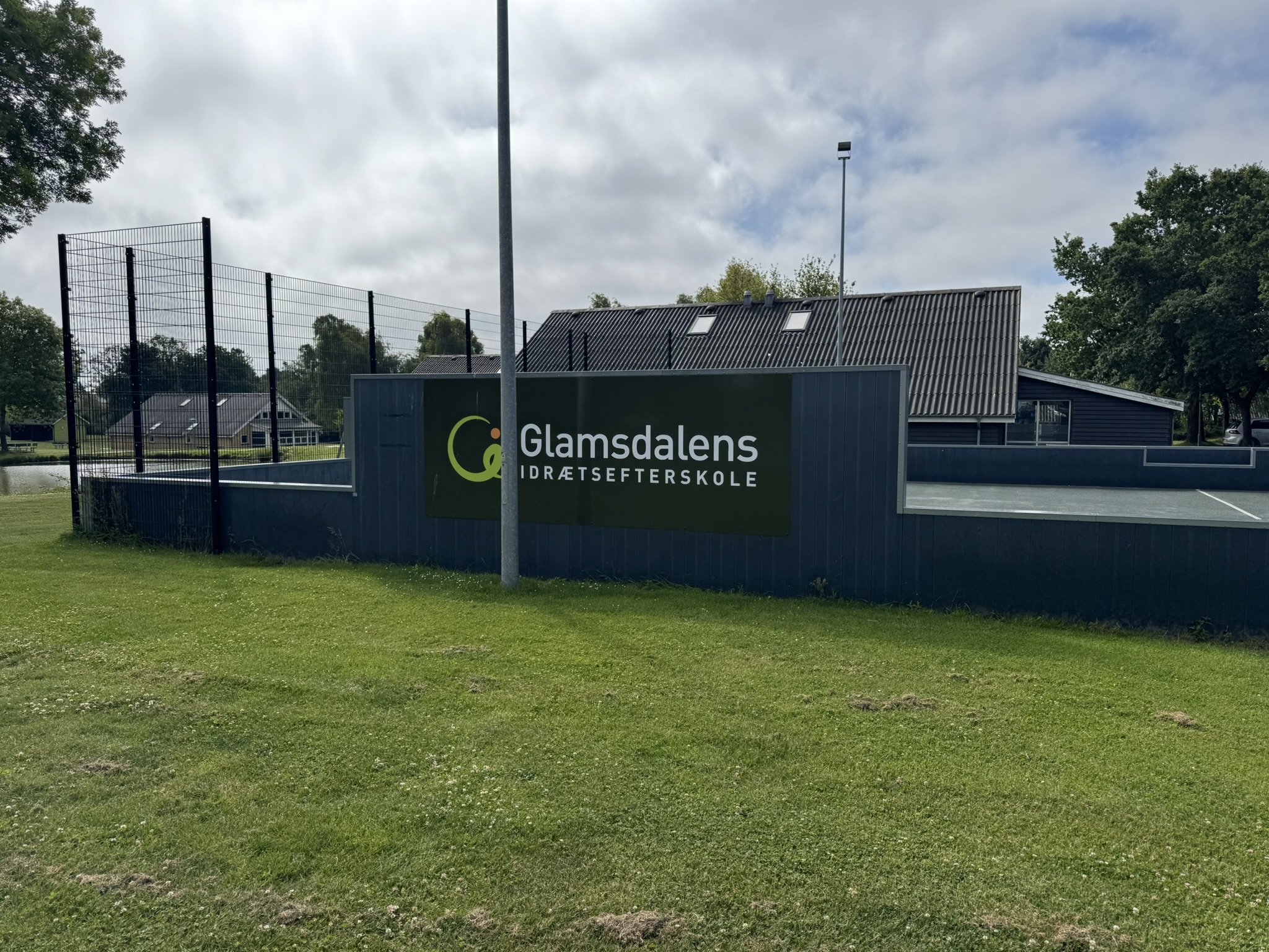 Skilt med teksten Glamsdalens Idrætsefterskole foran en sportsbane med en høje hegn og vandrestier, omgivet af grønne træer og huse under en overskyet himmel.
