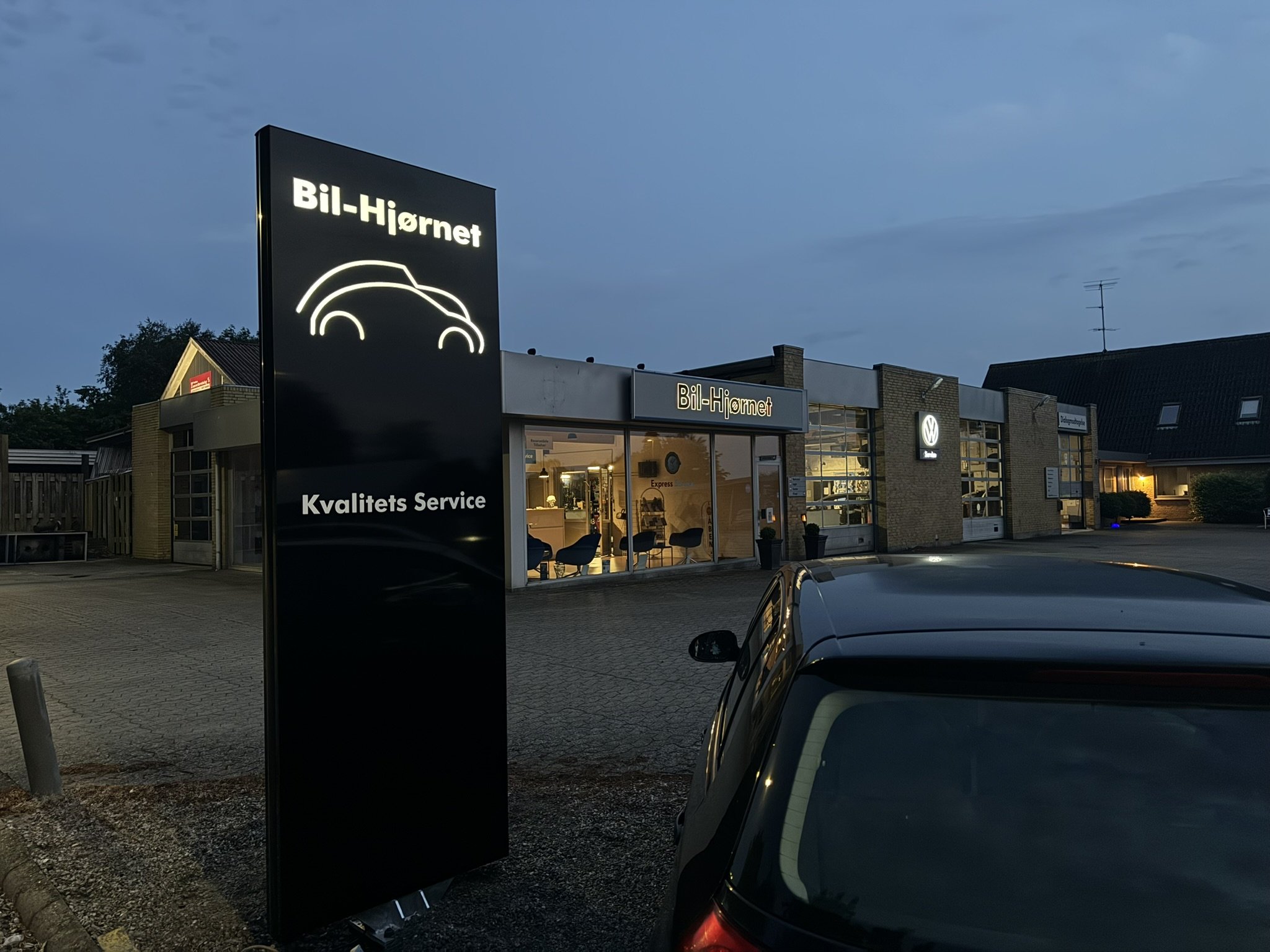 Et bilværksted ved aftenstid med belysning, der viser skilt med navnet 'Bil-Hjørnet' og logo, samt indgang til værkstedet hvor der er stole og vinduer, og biler parkeret udenfor.