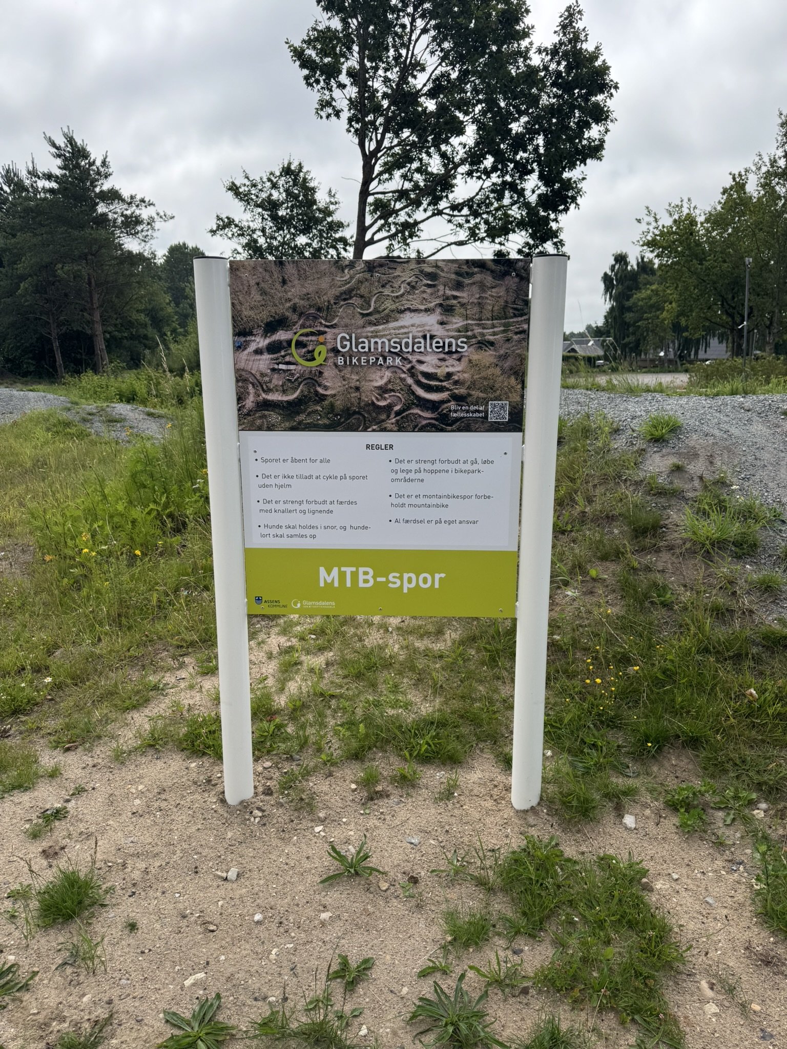 Informationsskilt ved MTB-spor i Glamsdalen Bikepark, omgivet af græs, sand og træer.