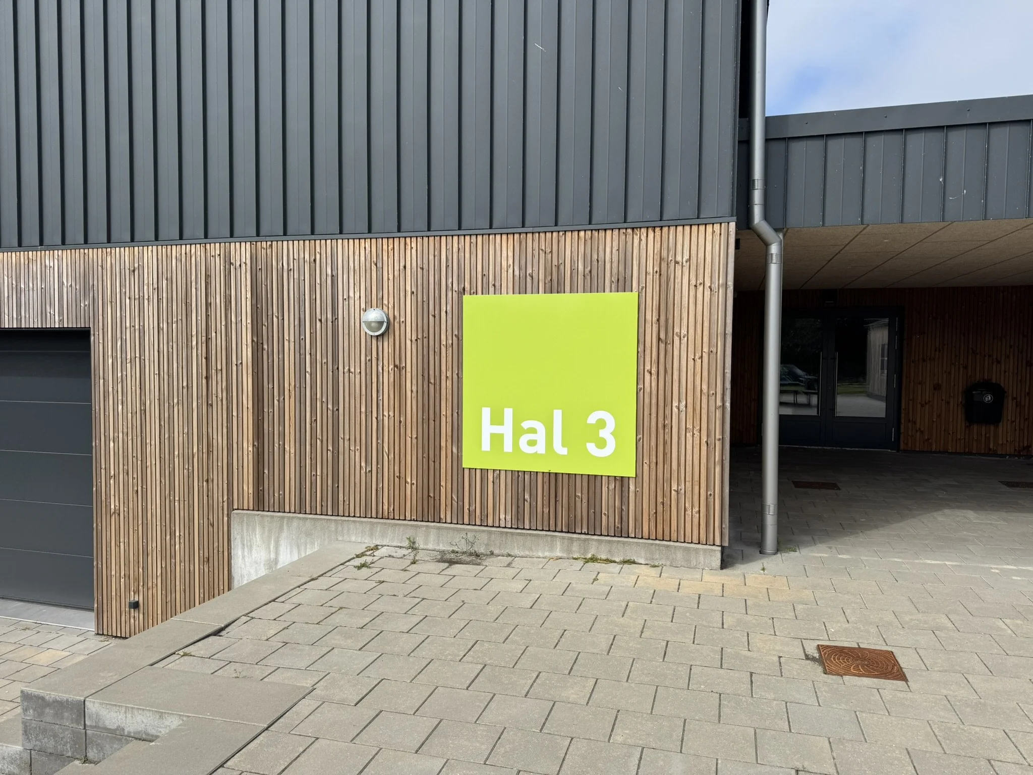Fasadet på en moderne bygning med to typer af beklædning: øverst grå metalpaneler og nederst naturligt træ. Der er en grøn skilt med teksten 'Hal 3', en udendørslampe og en nedløbsrør. Gade med brosten og fortov foran bygningen.