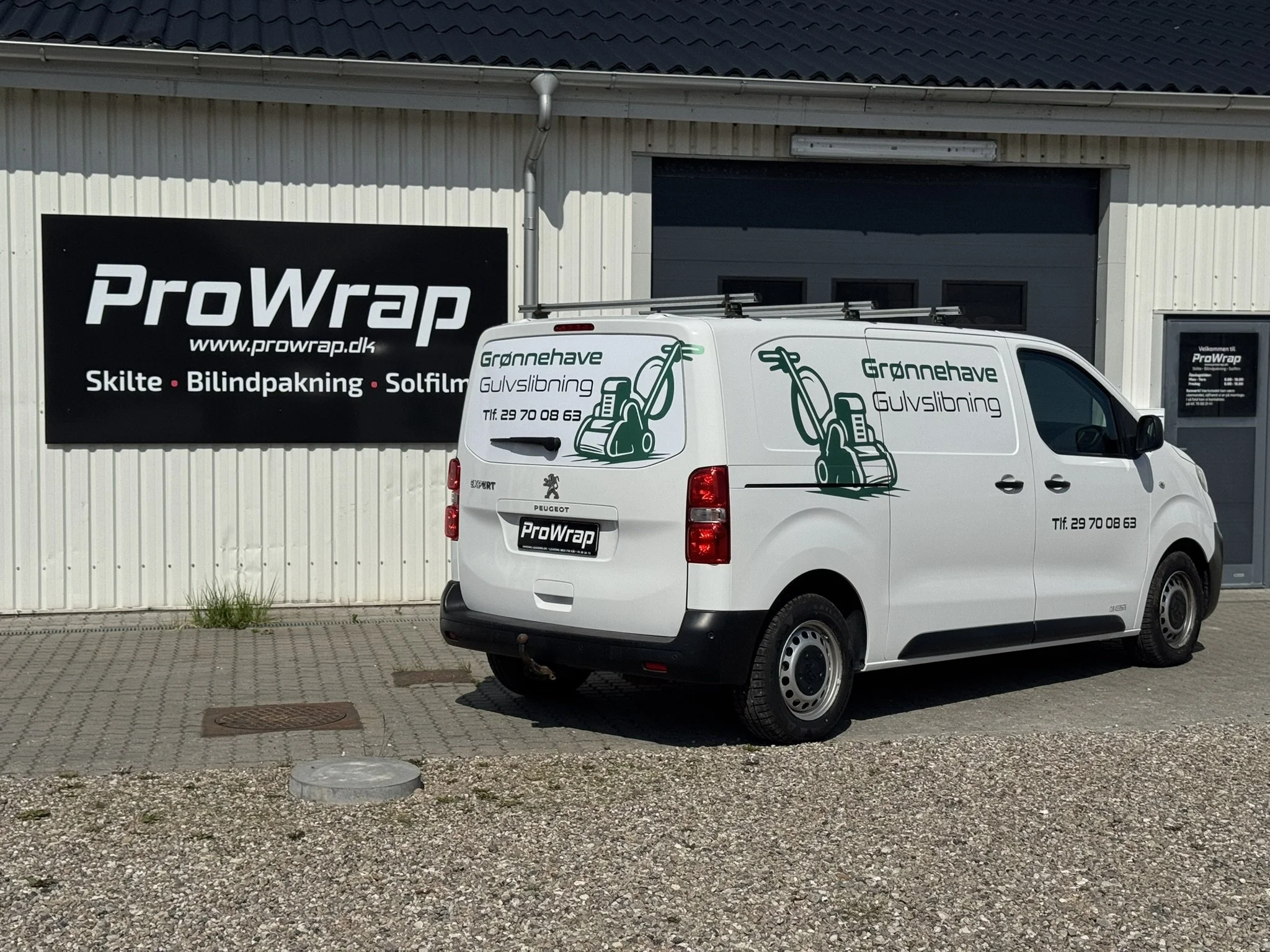 En hvid varevogn med grønne illustrationer og tekst, parkeret foran en bygning med skilt, der reklamerer for ProWrap og relaterede tjenester. Varevognen har logo og kontaktoplysninger.