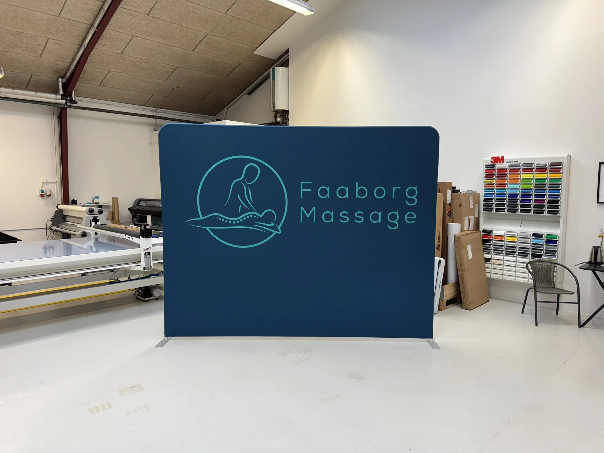 En indendørs arbejdsplads med en blå skærm, der har teksten 'Fåborg Massage', en printer, farve- og materialeprøver samt nogle papkasser.