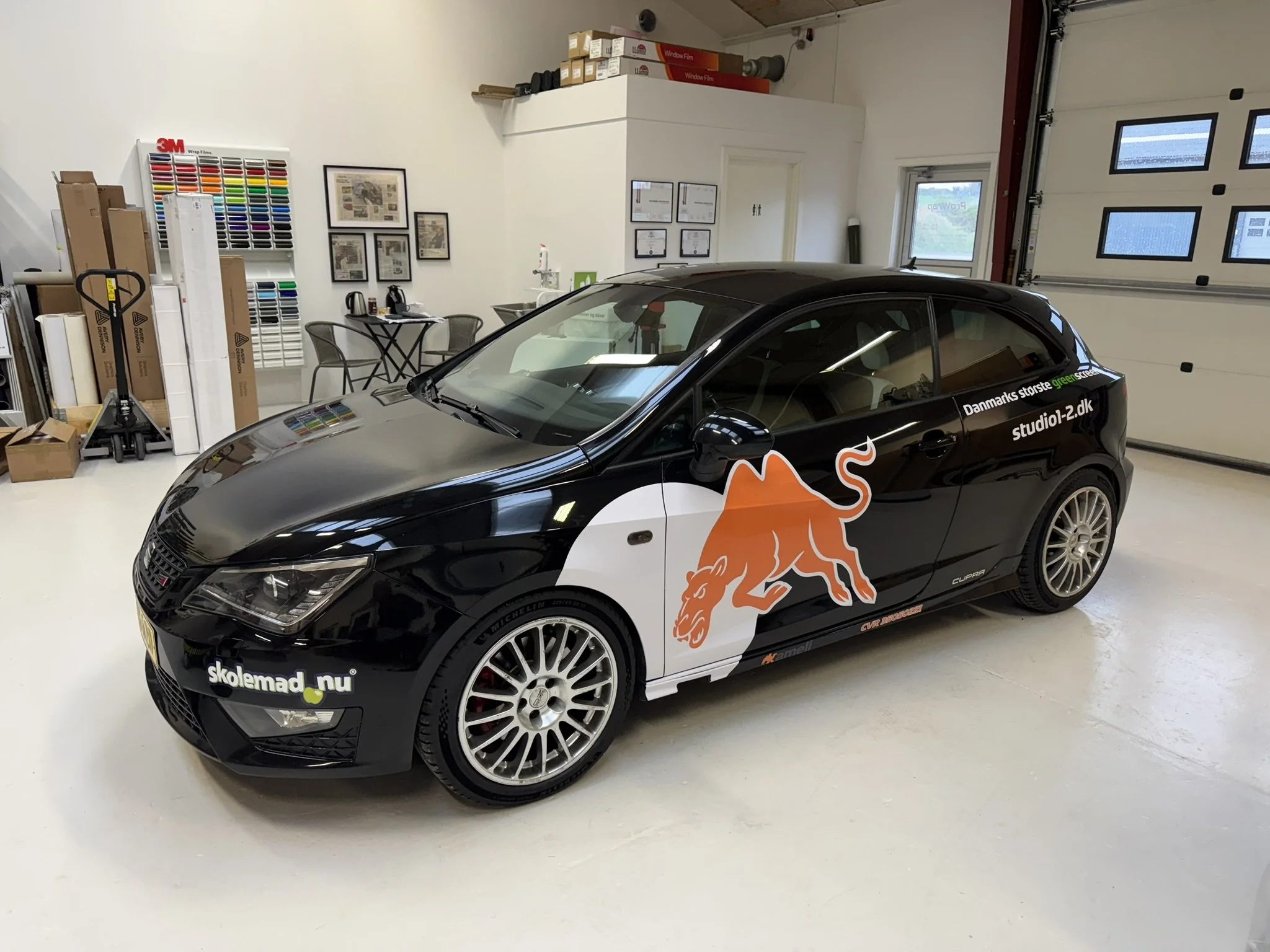 En sort bil med en orange løvegrafik og tekst, der står 'skolemad.nu', parkeret i et garage med vinduer og certificeringsdokumenter på væggen.
