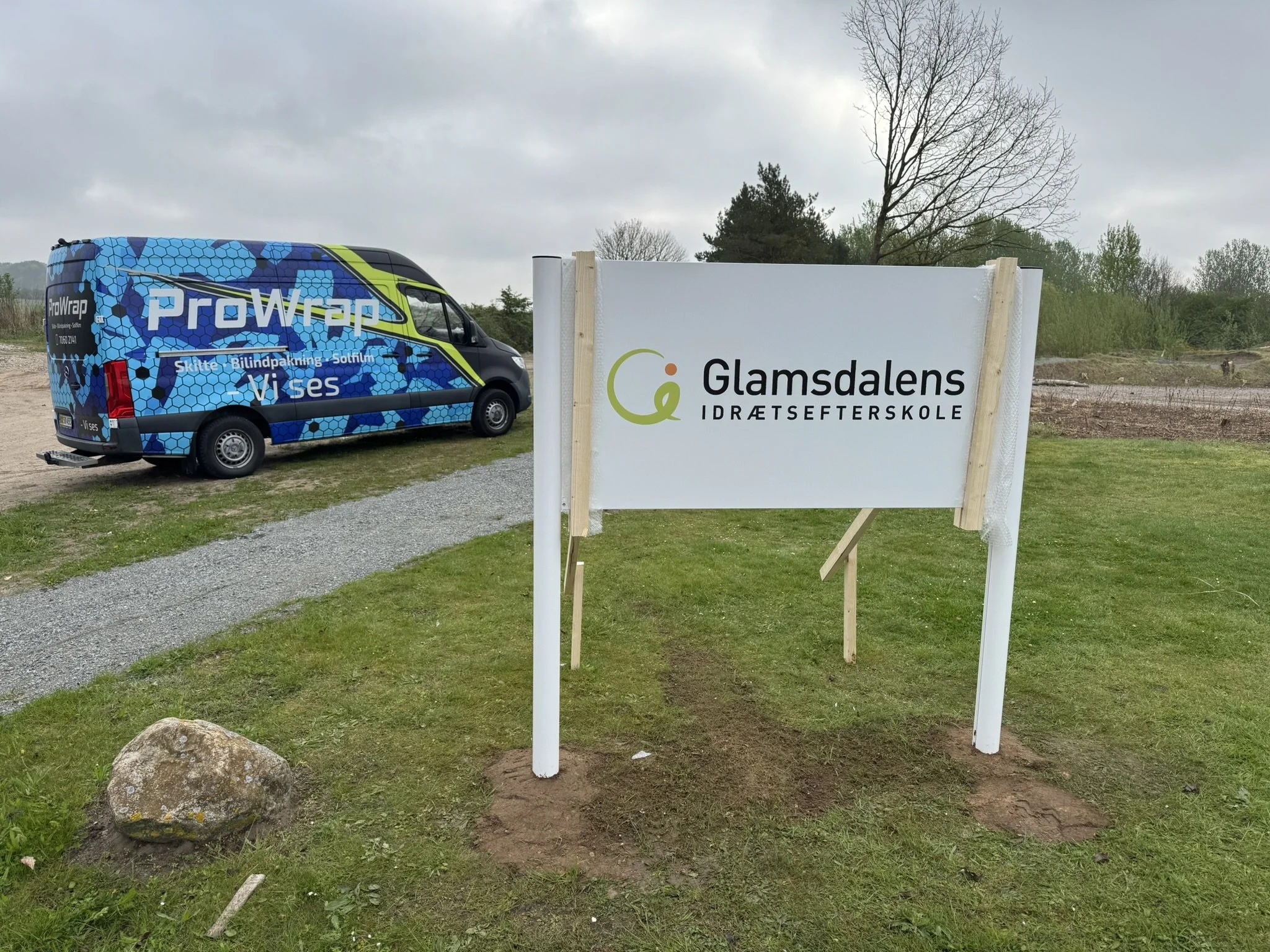 En markerplads med en reklamebil til højre, der har blå, grøn og sort front, inklusive teksten 'ProWrap' og 'Skilte - Bilindpakning - Solfilm - Visses'. Der står også et skilt til venstre med teksten 'Glamdals Idrætsefterskole'. Baggrund med nogle træer og en overvejende overskyet himmel.