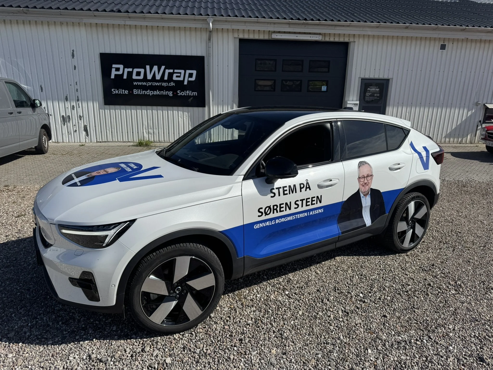 Billede af en hvid bil med reklame for valgkampagne med en persons ansigt og tekst, parkeret foran en industribygning med skilte for Prowrap, der tilbyder skilte, bilindpakning og solfilm.