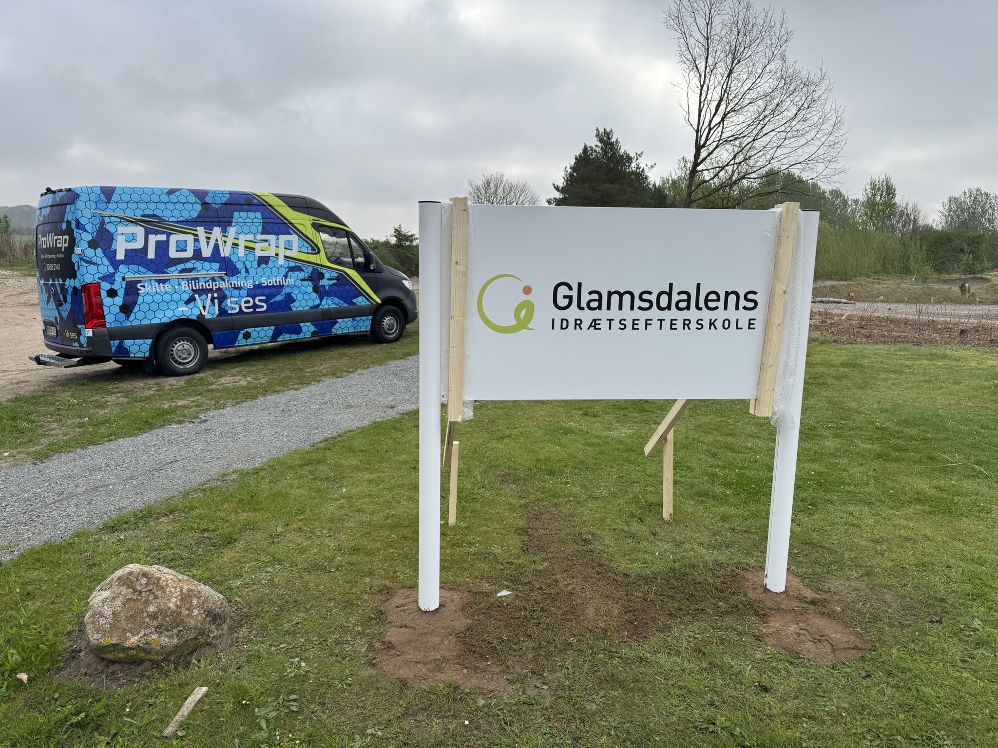 En blå reklamevaretvogn med logo og tekst, parkeret ved en byggeplads eller udendørs område med græs og træer, og en skilt med teksten "Glamdals Sports School".