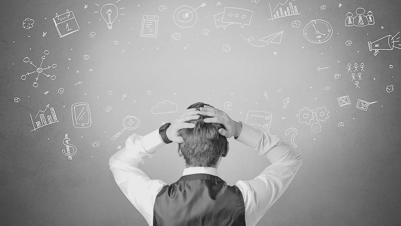 A man with short hair, dressed in a suit, stands with his back to the camera, holding his head with both hands. He is in front of a gray wall with various white chalk drawings of charts, graphs, gears, a clock, a light bulb, a calendar, and other business-related icons.