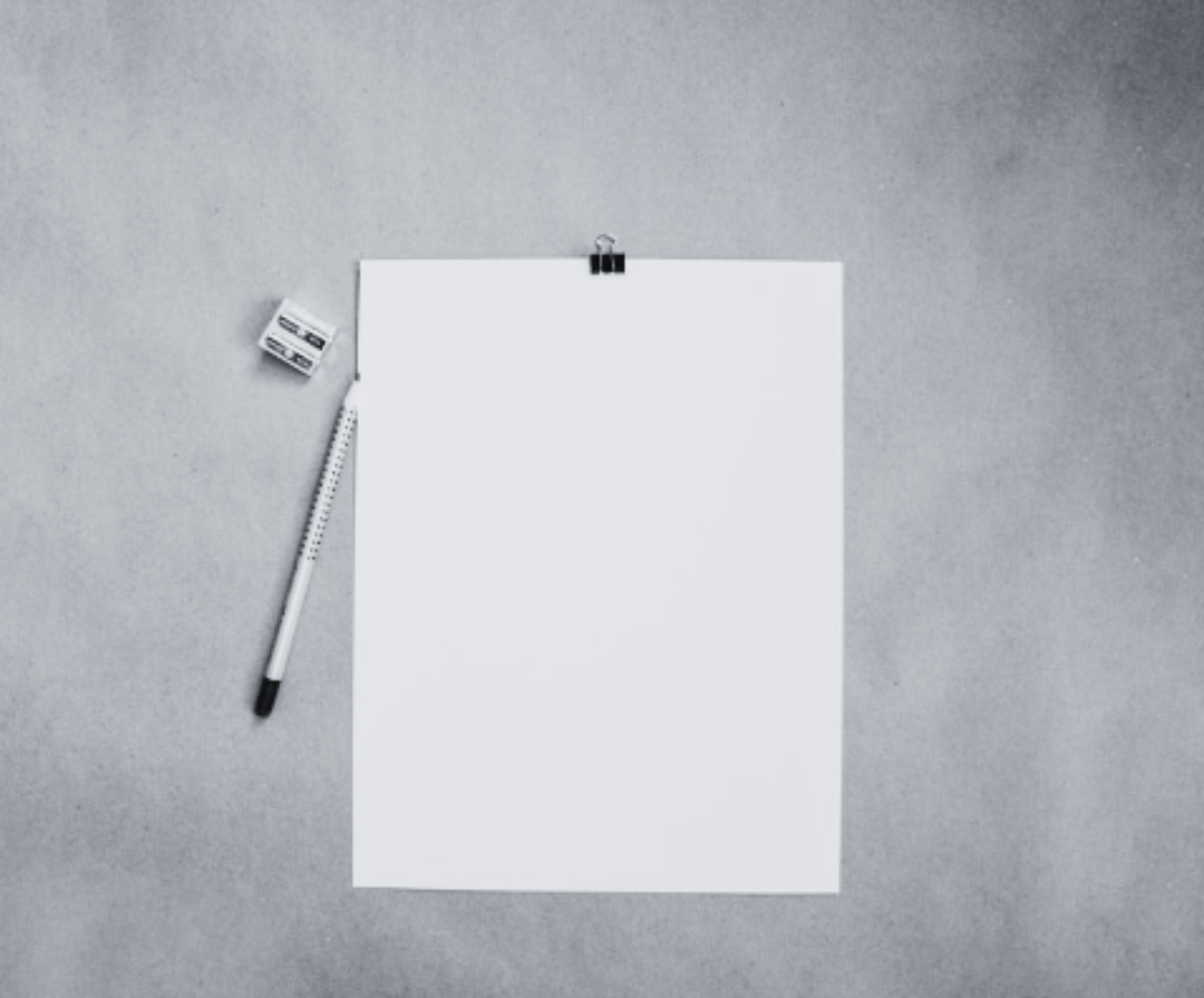 A blank white sheet of paper attached to a gray background with a black clip, a black and white pen, and a small white eraser nearby.