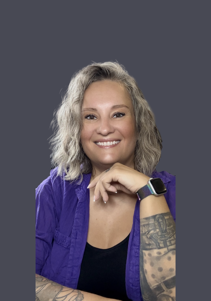 A woman with shoulder-length curly gray hair, smiling and wearing a black top with a purple shirt and a smartwatch, showing tattoos on her left arm, against a plain gray background.