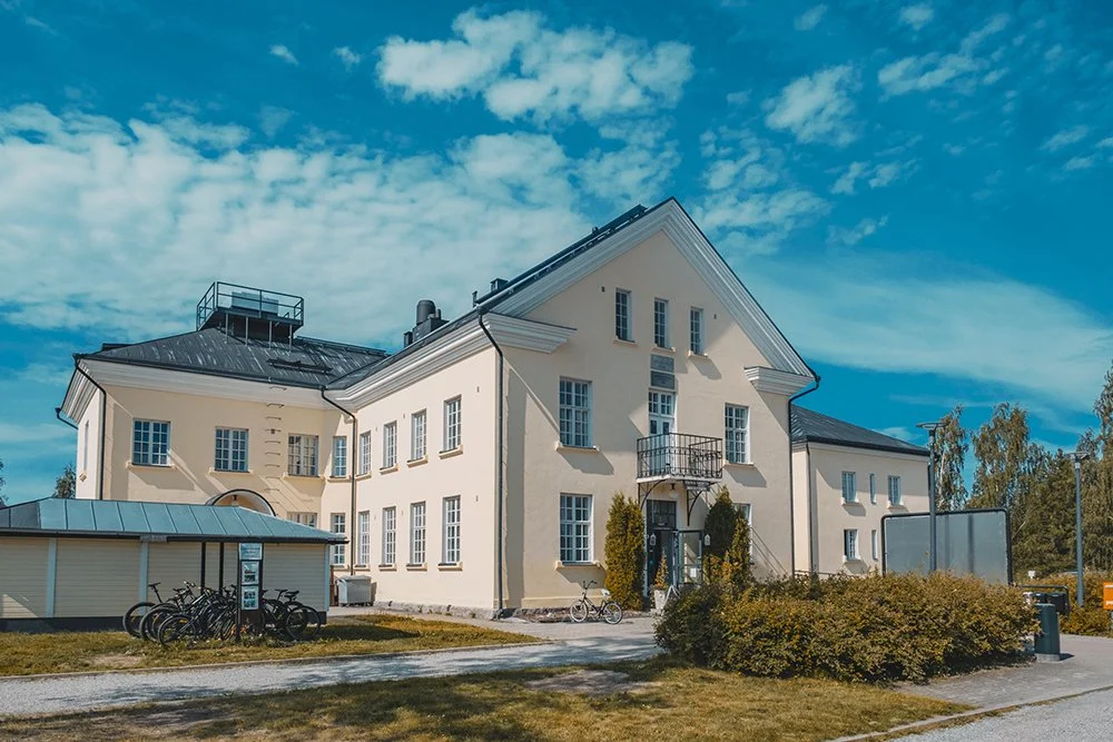 Large beige building with multiple windows, surrounded by greenery, and bicycles parked in front, under a partly cloudy blue sky.