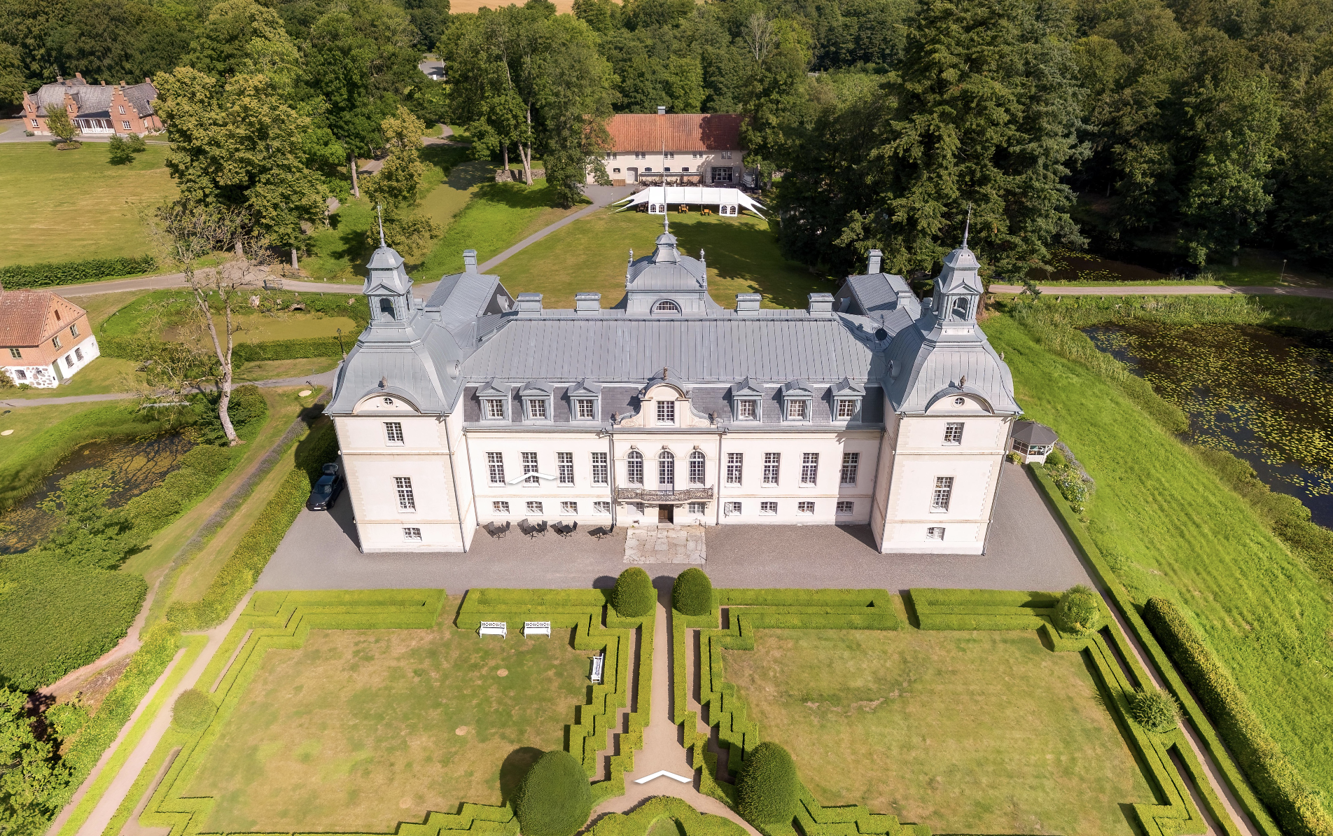 Aerial view of a historical European chateau with manicured gardens, surrounded by lush green trees and pathways. The building features ornate architectural details and multiple dormer windows. The landscape includes a pond, lawns, and smaller buildings nearby.