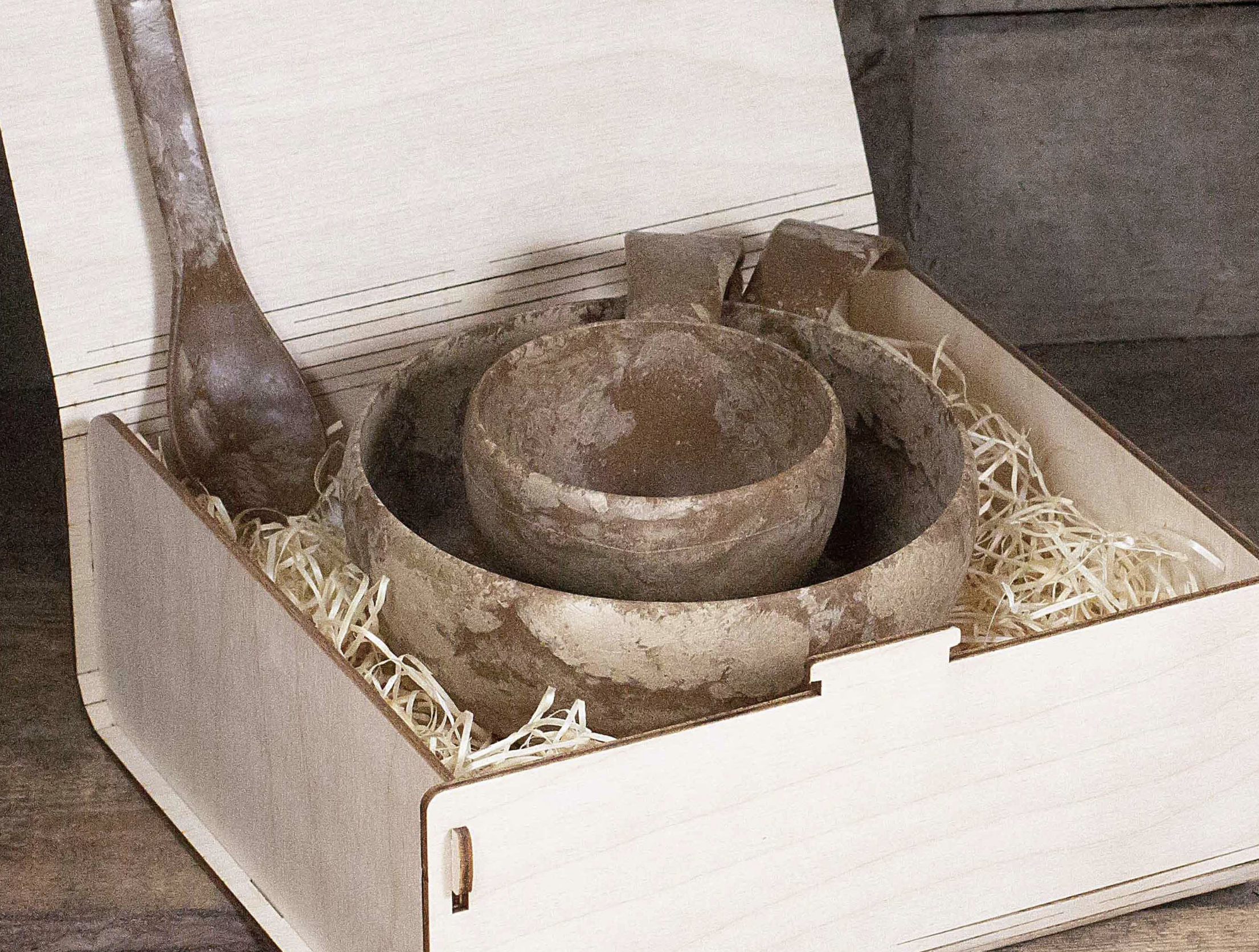 A wooden box containing a set of brown earthenware bowls and a spoon, surrounded by straw packing material. The lid of the box is partially open.