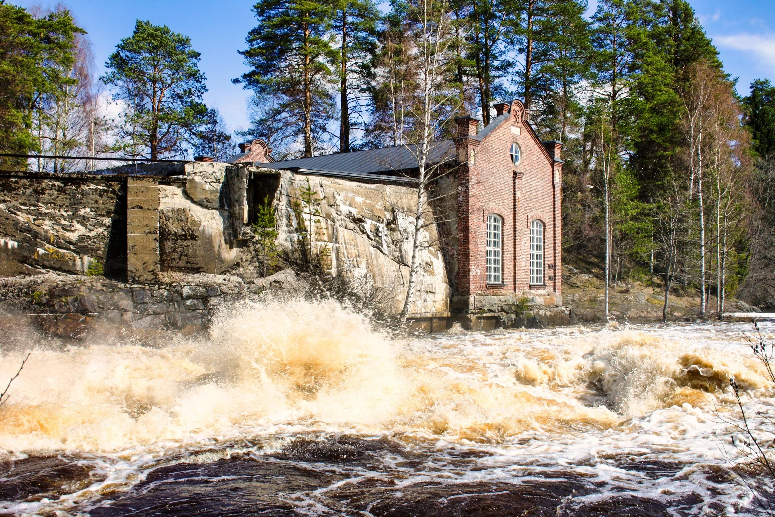 Rushing river water splashing against rocks and an old brick building with large windows, surrounded by trees.