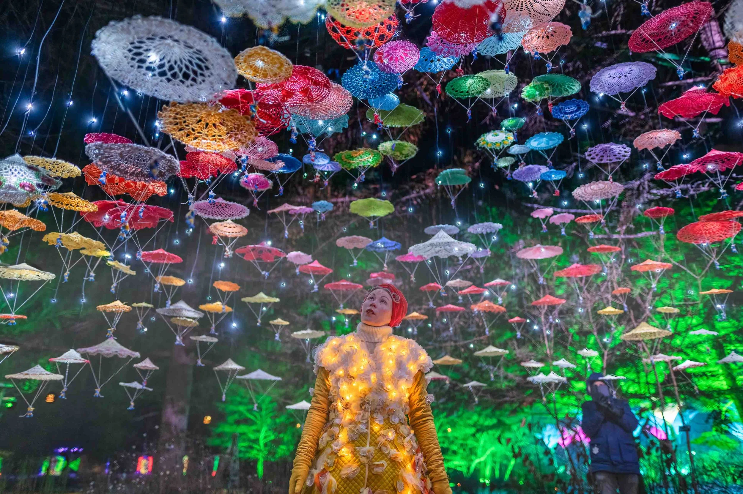 Person in illuminated costume looking up at colorful crochet discs hanging from garden ceiling.