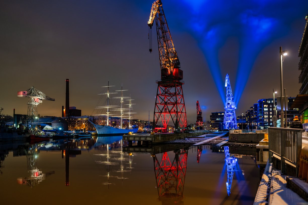 Nighttime harbor scene with illuminated cranes and a sailboat, city skyline in the background, reflections in calm water.