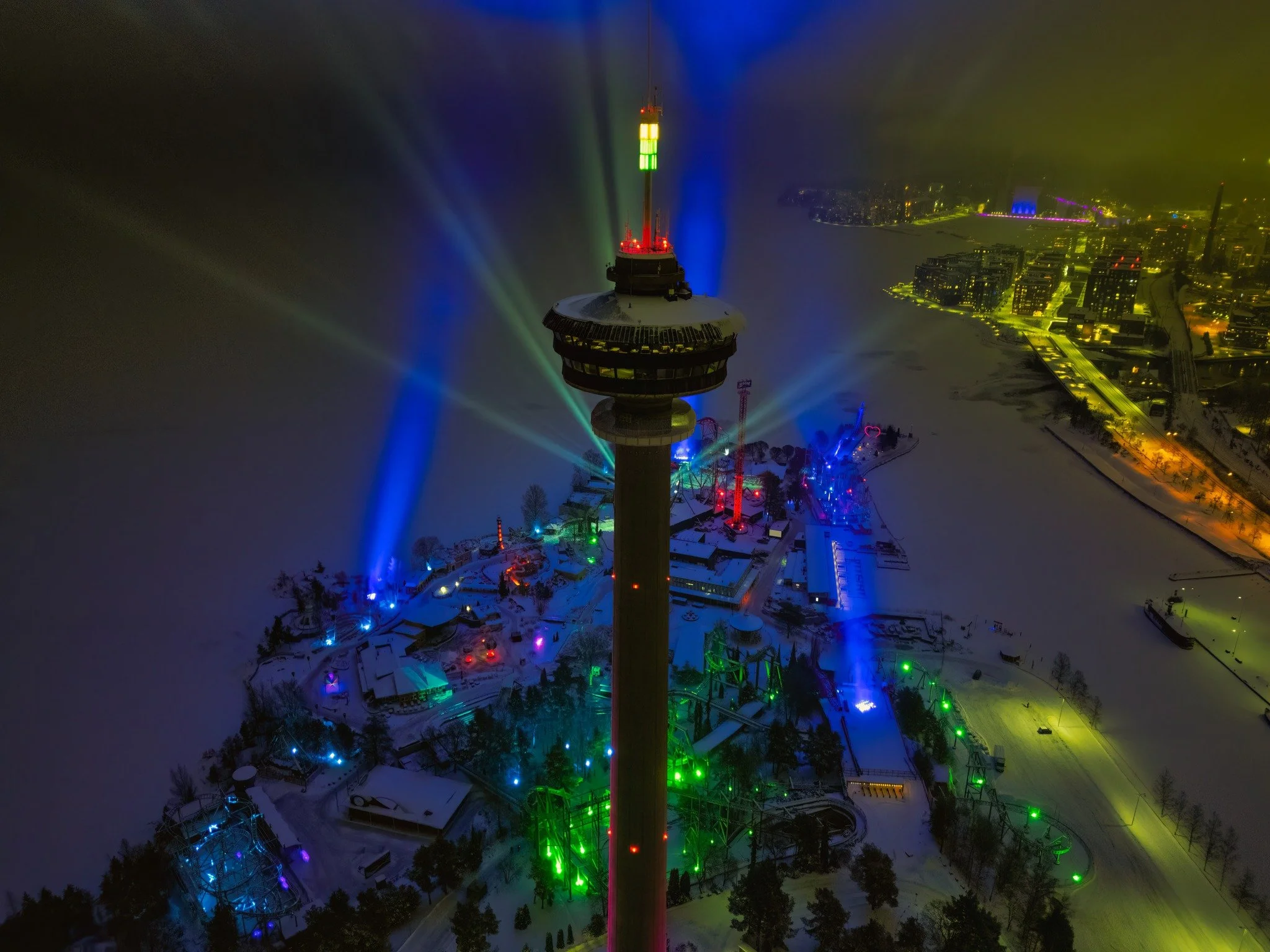 Aerial night view of an illuminated cityscape with a tall tower at the center. The surrounding area features colorful lights in blue, green, and red, possibly from an amusement park. Snow covers the ground, creating a winter landscape.