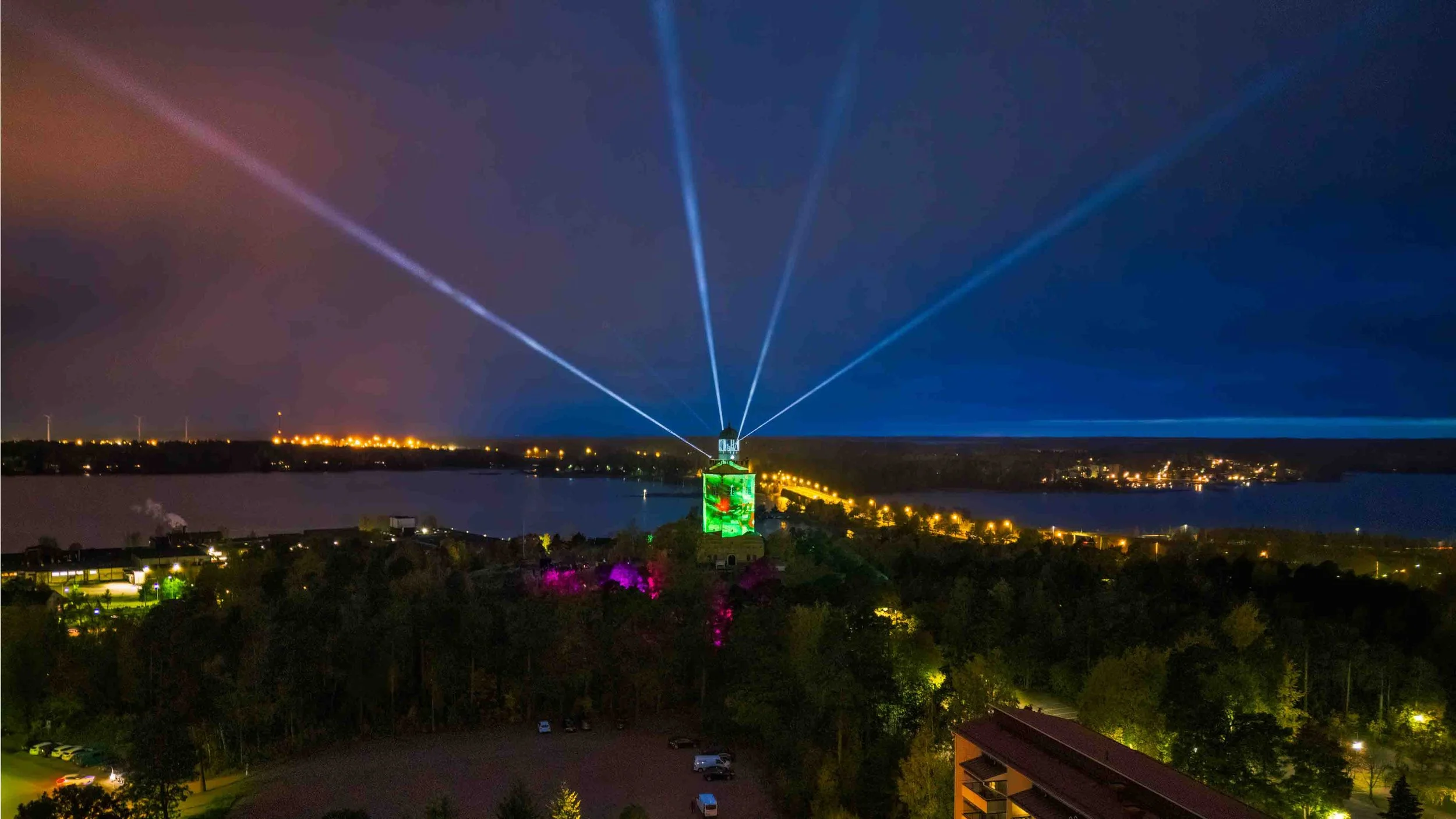 Nighttime scene with illuminated tower displaying colorful lights and beams, surrounded by a lit-up town and water, under a dark sky.