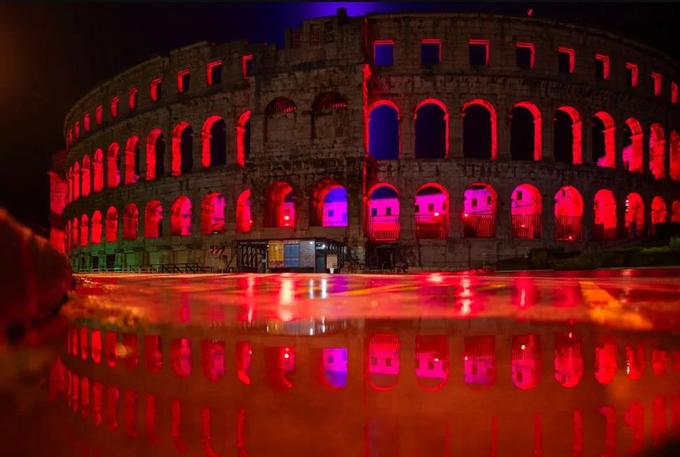 Night view of the Roman Colosseum illuminated with pink, orange, and purple lights, with its reflection visible in a puddle on the ground.